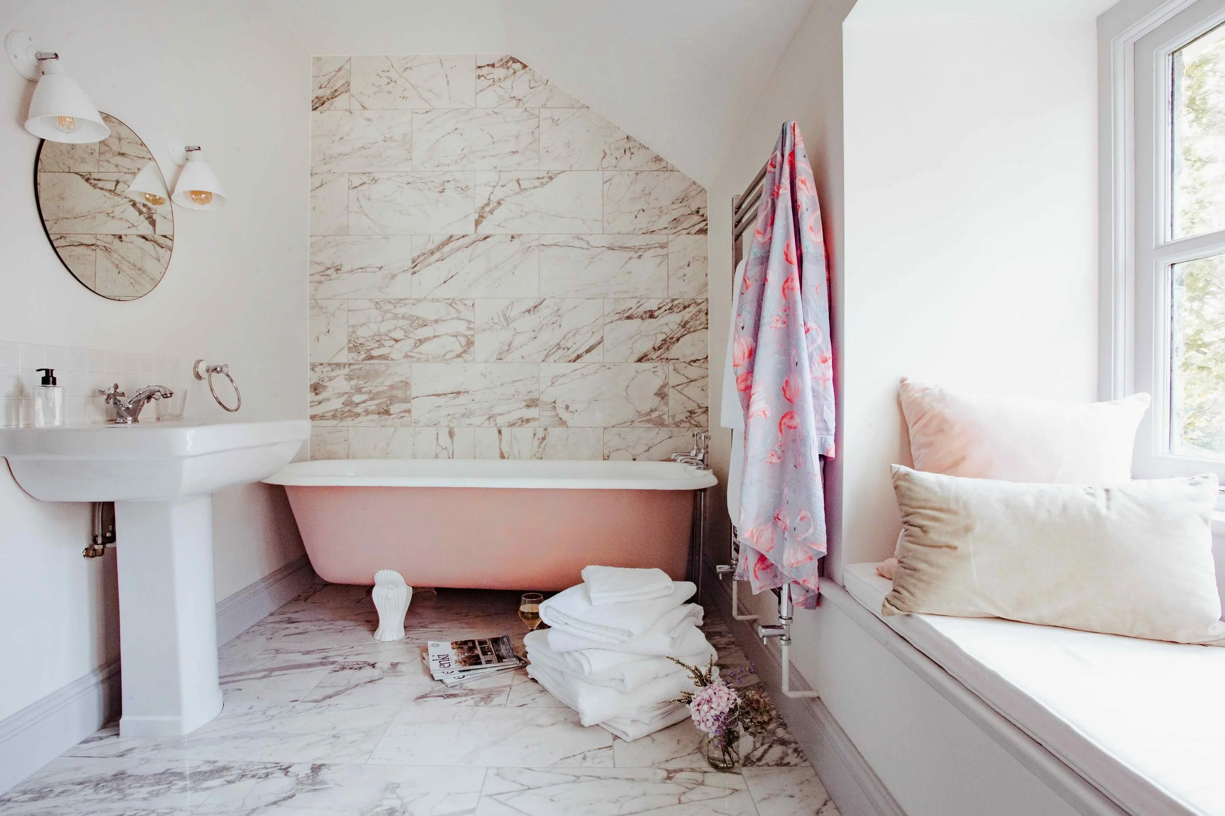 Bathroom with a pink clawfoot bathtub, white pedestal sink, marble tiles on the wall and floor, a window seat with pillows, folded towels, a magazine, and flowers.