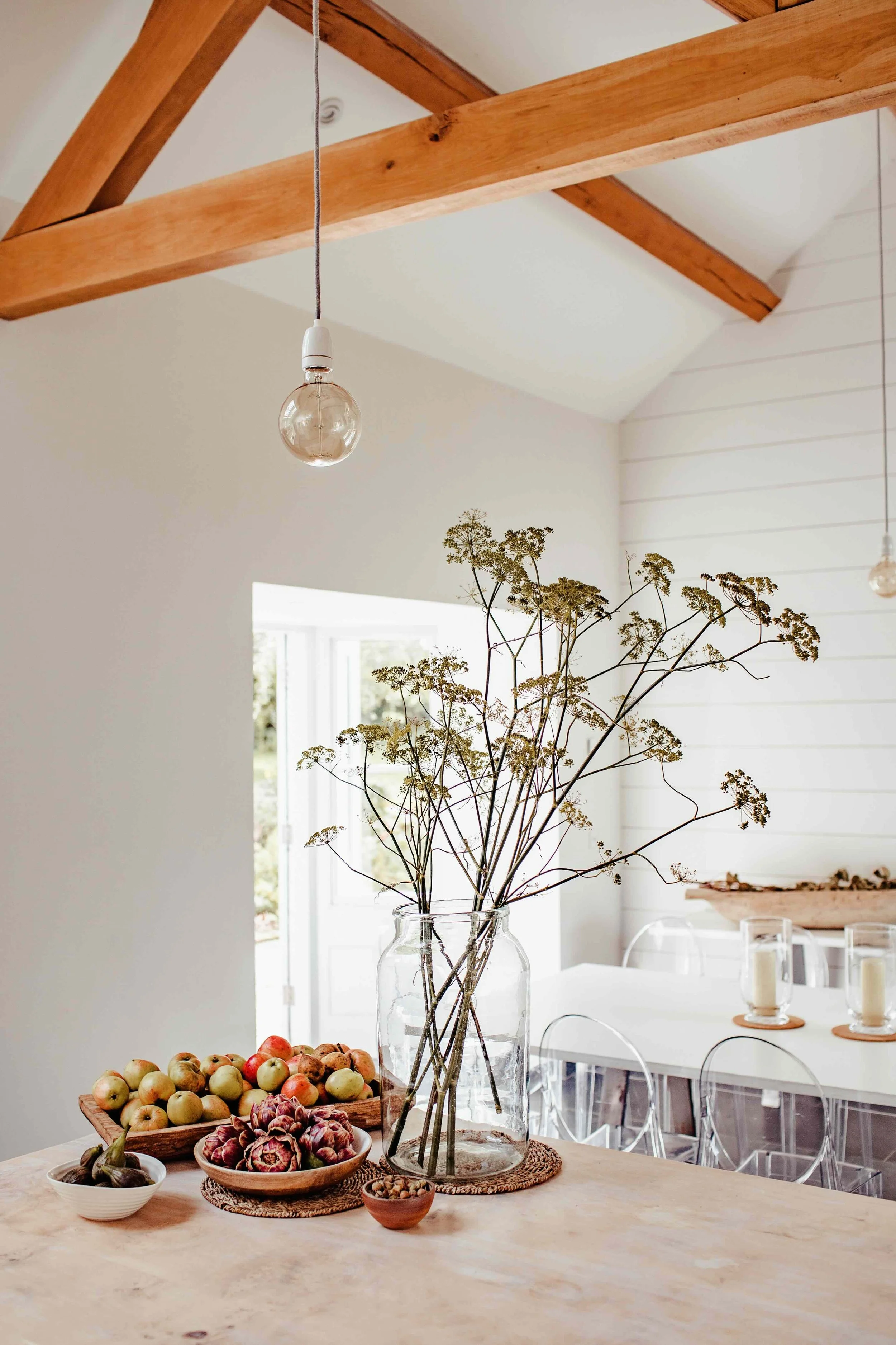 Interior view of a bright, minimalistic dining area with a wooden table, a large glass vase holding dried branches, and a variety of fresh fruits including apples, figs, and nuts on the table. Light from a nearby window illuminates the space, with pe