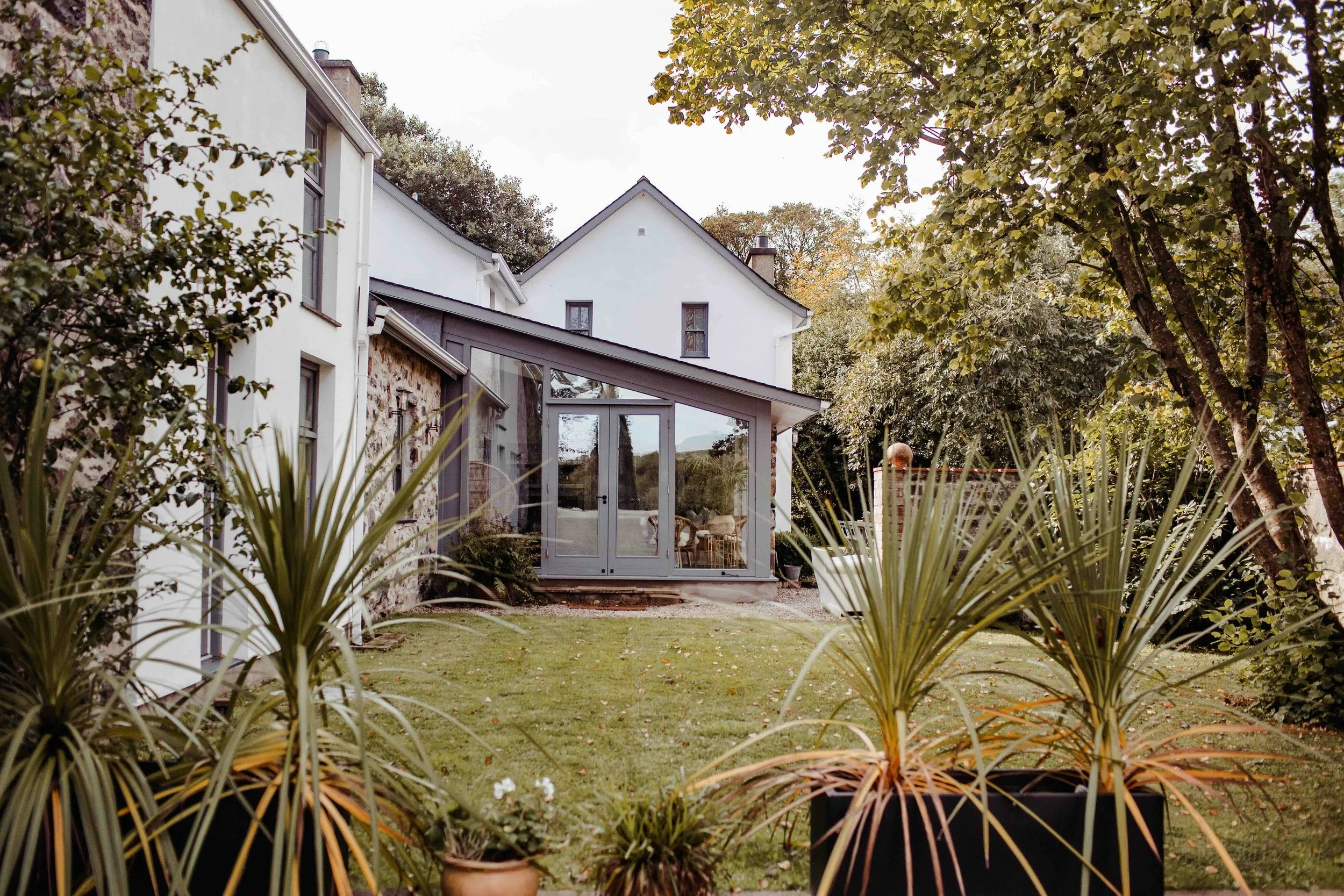 A backyard garden with green grass, potted plants in the foreground, trees on the right, and a modern house with a glass extension at the back.