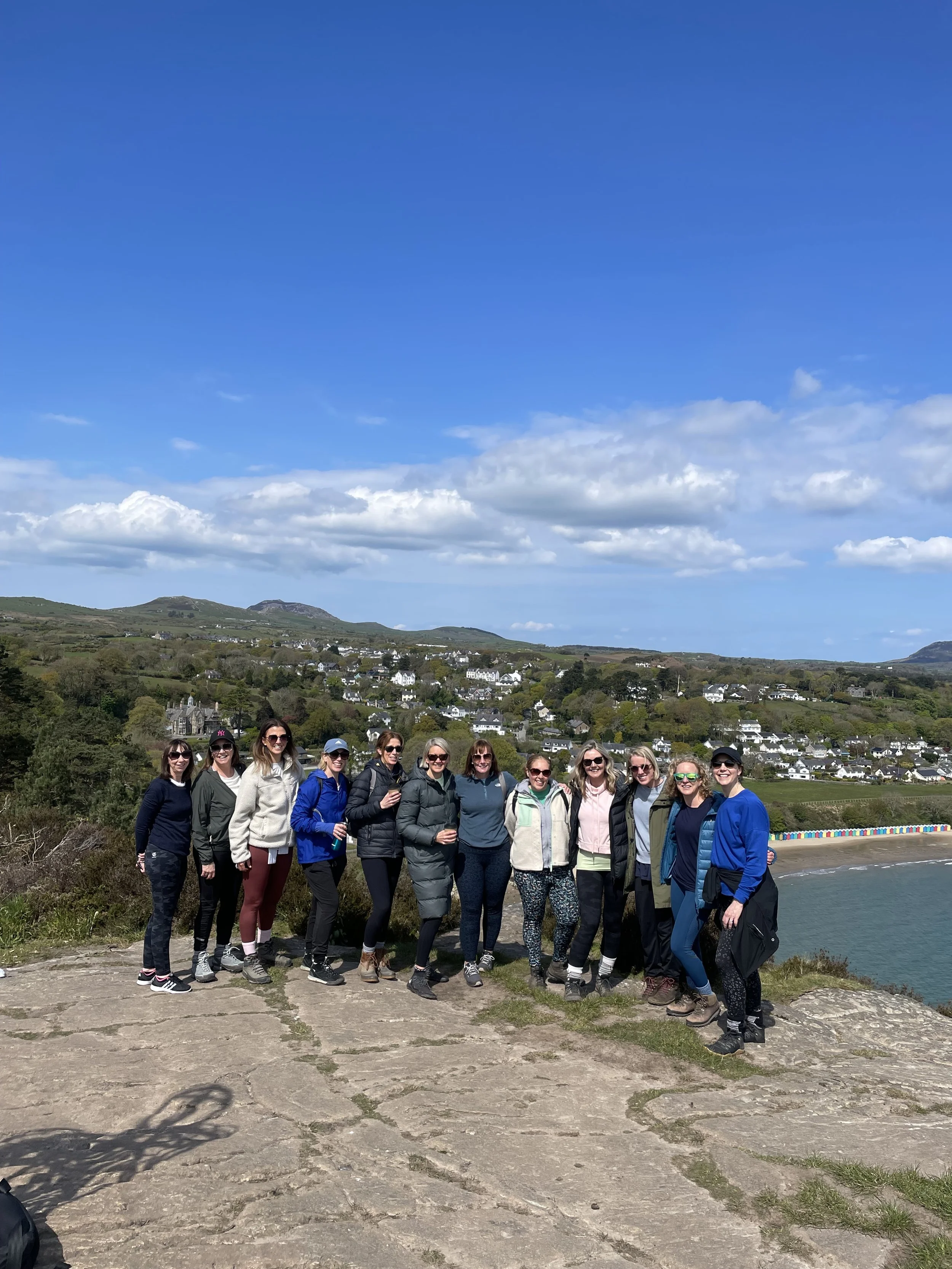 A group of 12 women standing outdoors on a hilltop with a scenic view of the coastline, houses on the hillside, and a bright blue sky with scattered clouds.