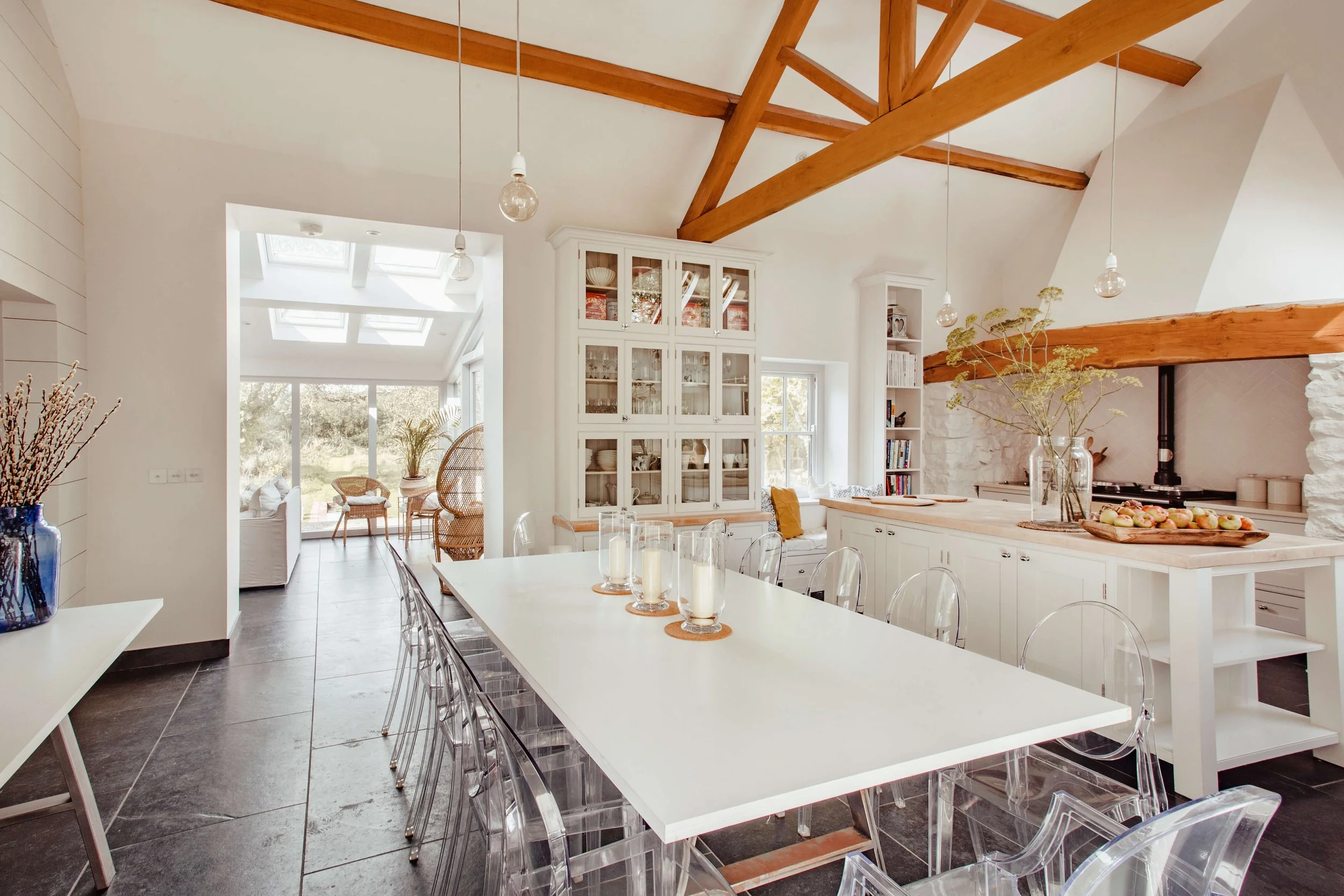 Bright open-concept kitchen with white cabinetry, a large white dining table with transparent chairs, a wooden beam ceiling, and a cozy sitting area with wicker chairs in the background, decorated with candles and vases with flowers.