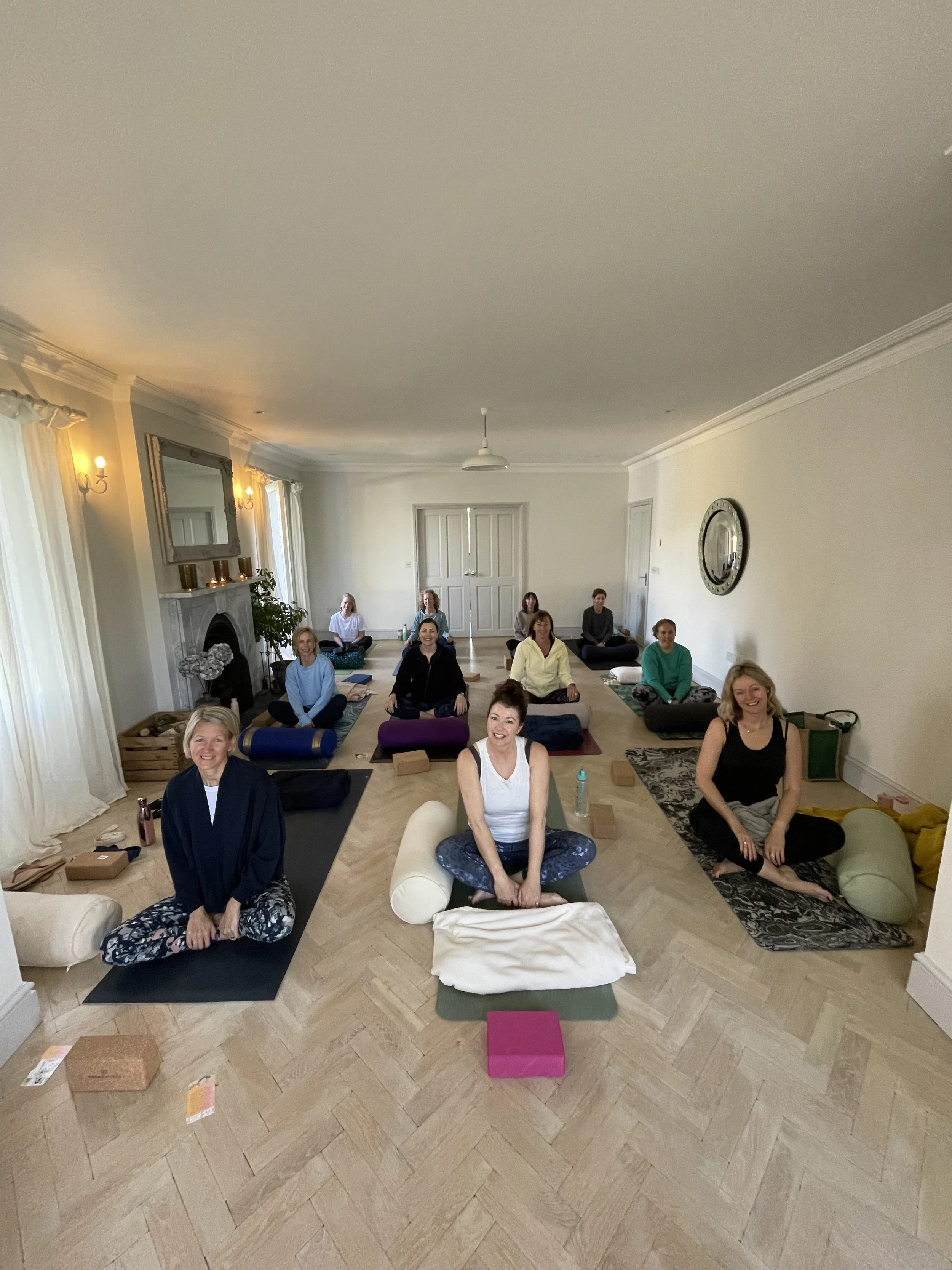 A group of women participating in a yoga or meditation class in a spacious, well-lit living room with large windows, curtains, a mirror above the fireplace, and a clock on the wall.