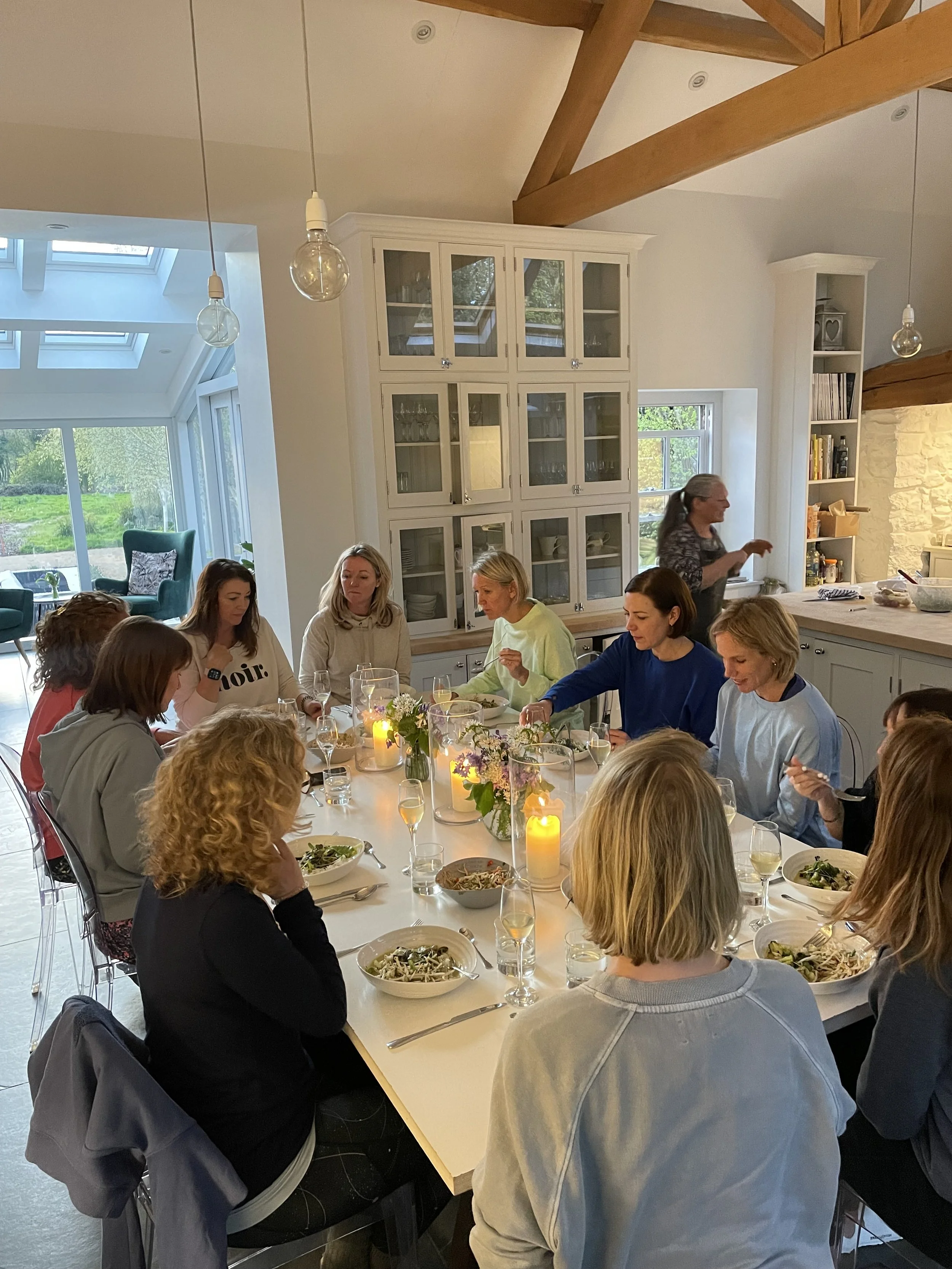 A group of women gathered around a dining table enjoying a meal in a bright, modern kitchen with large windows and wooden beams.