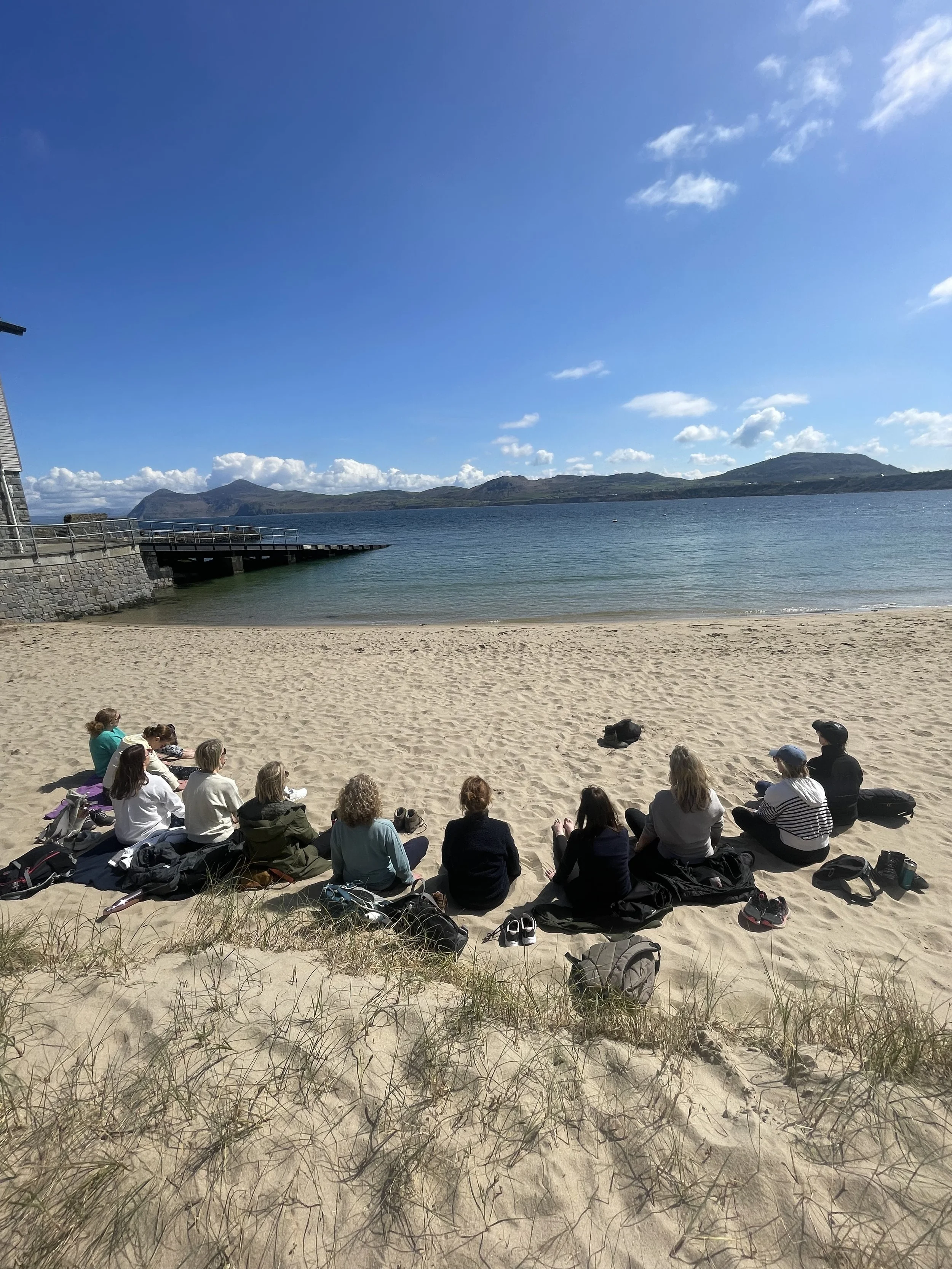 People sitting on the sandy beach facing the water and mountains in the distance under a blue sky with clouds.