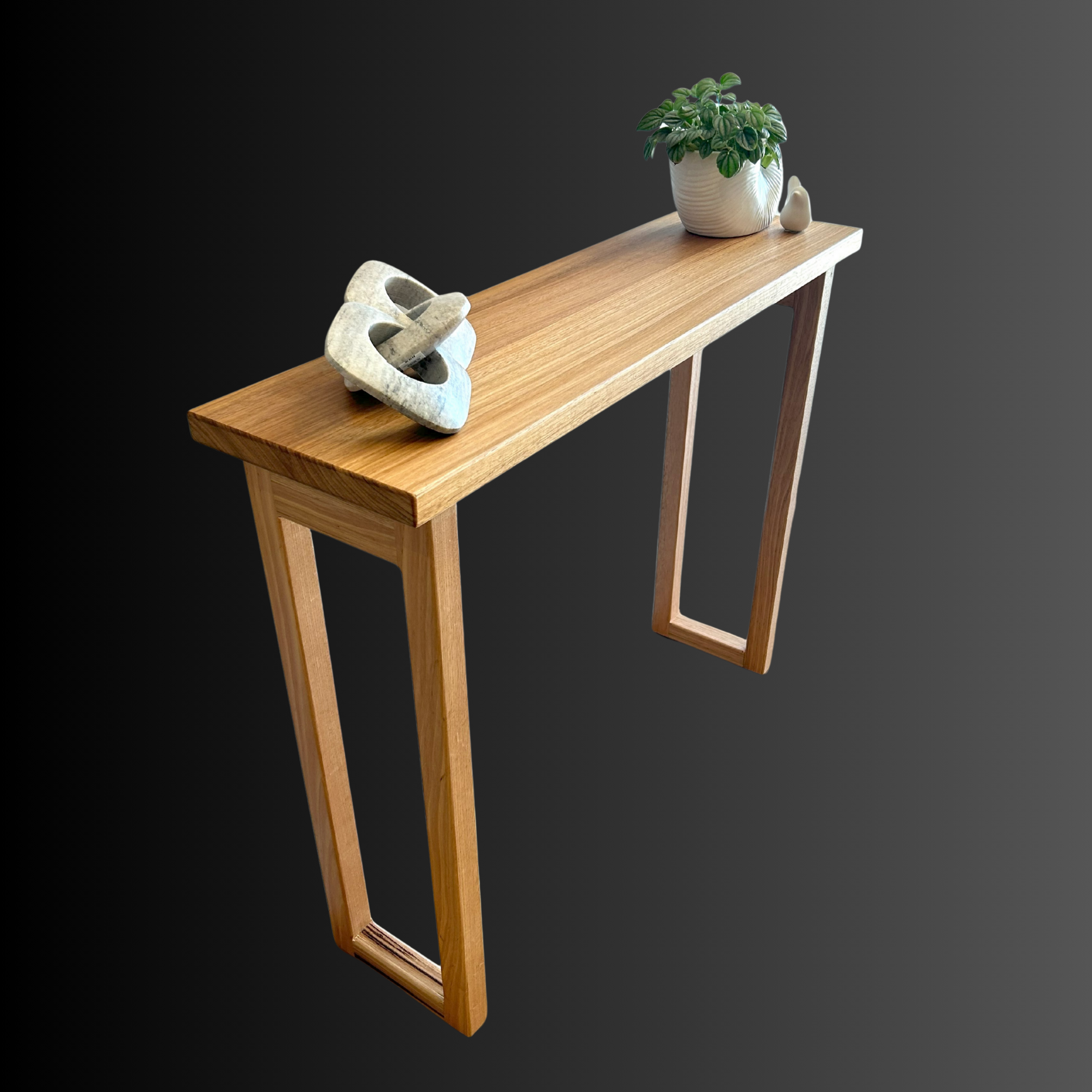 A wooden console table with a potted plant, two white decorative objects, and a sculptural chain on top, against a dark gray background.