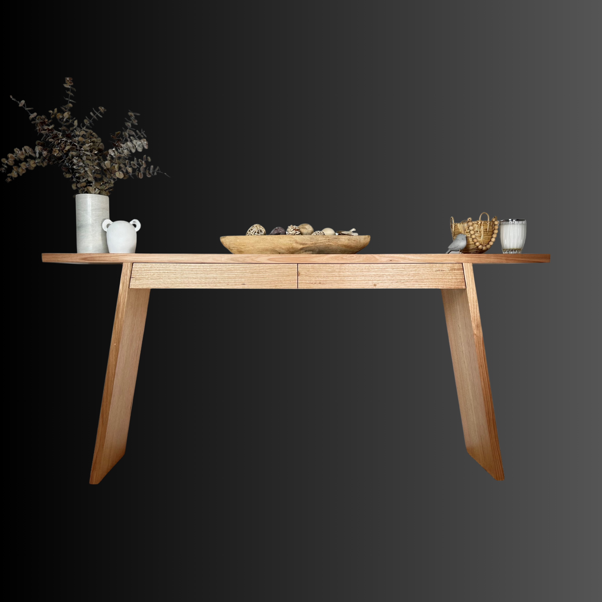 A wooden table with decorative items including a tall white vase with dried branches, a small white vase, a wooden bowl filled with decorative stones, a woven basket, and a glass of milk against a dark and gray background.