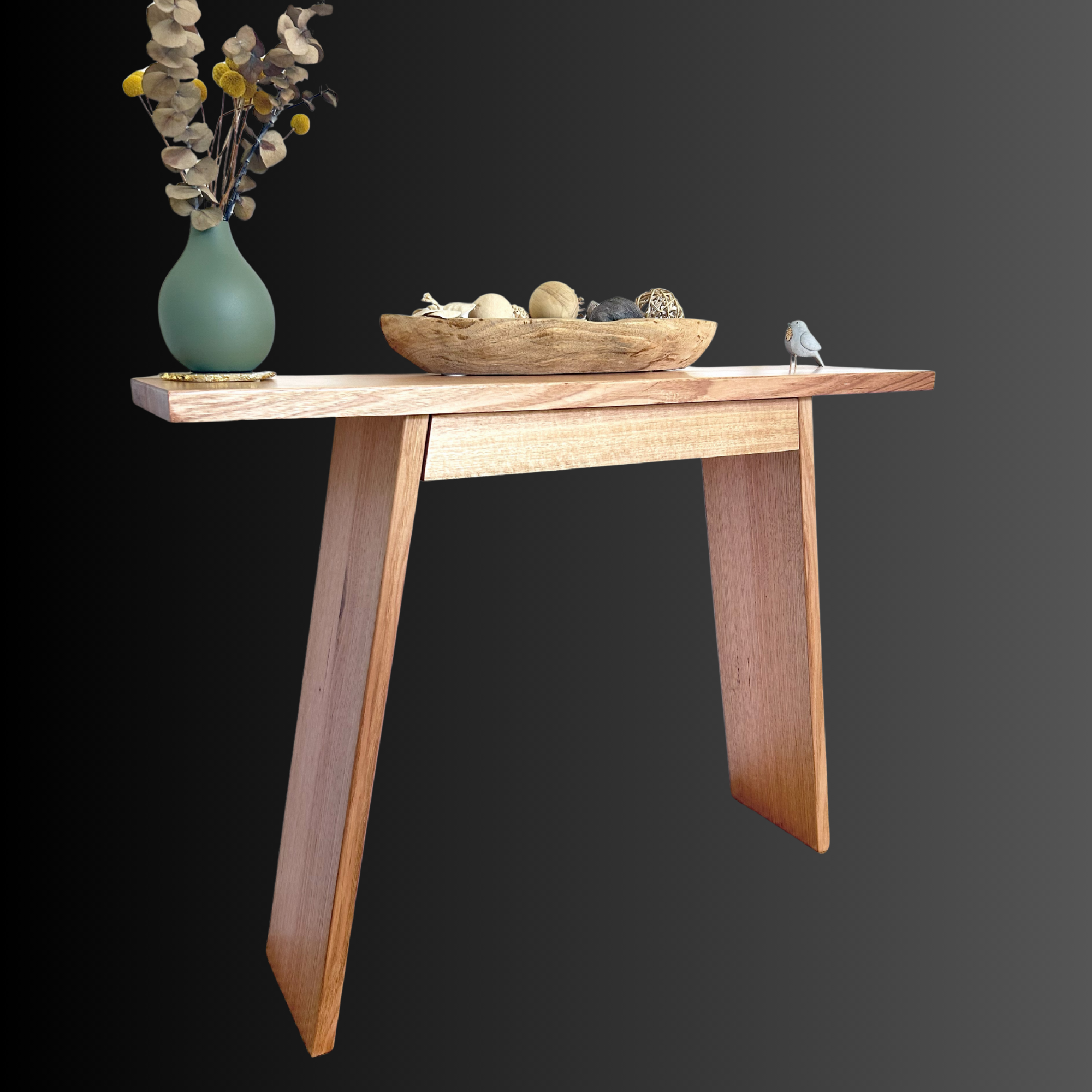 A wooden table with a tall green vase holding dried flowers, a wooden bowl filled with small decorative objects, and a small bird figurine on a black to gray gradient background.