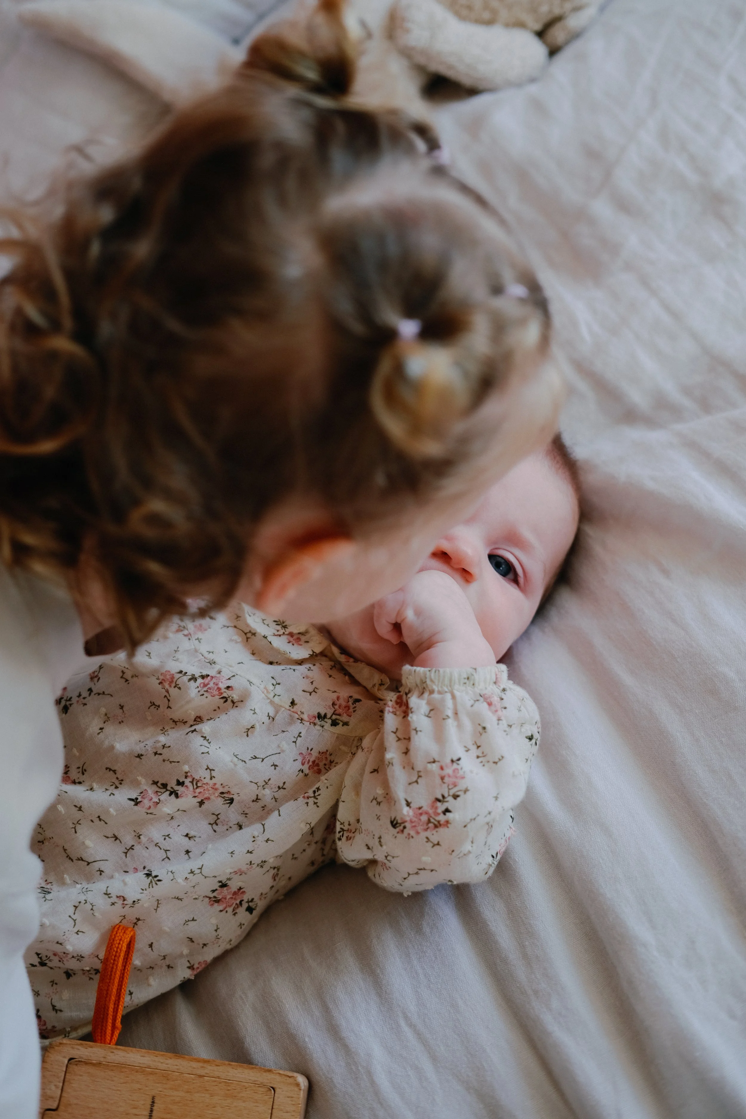 Une jeune femme aux cheveux bouclés voit un bébé allongé sur un lit avec des draps blancs. Le bébé porte une robe à fleurs et regarde la femme, tout en se portant la main à la bouche.