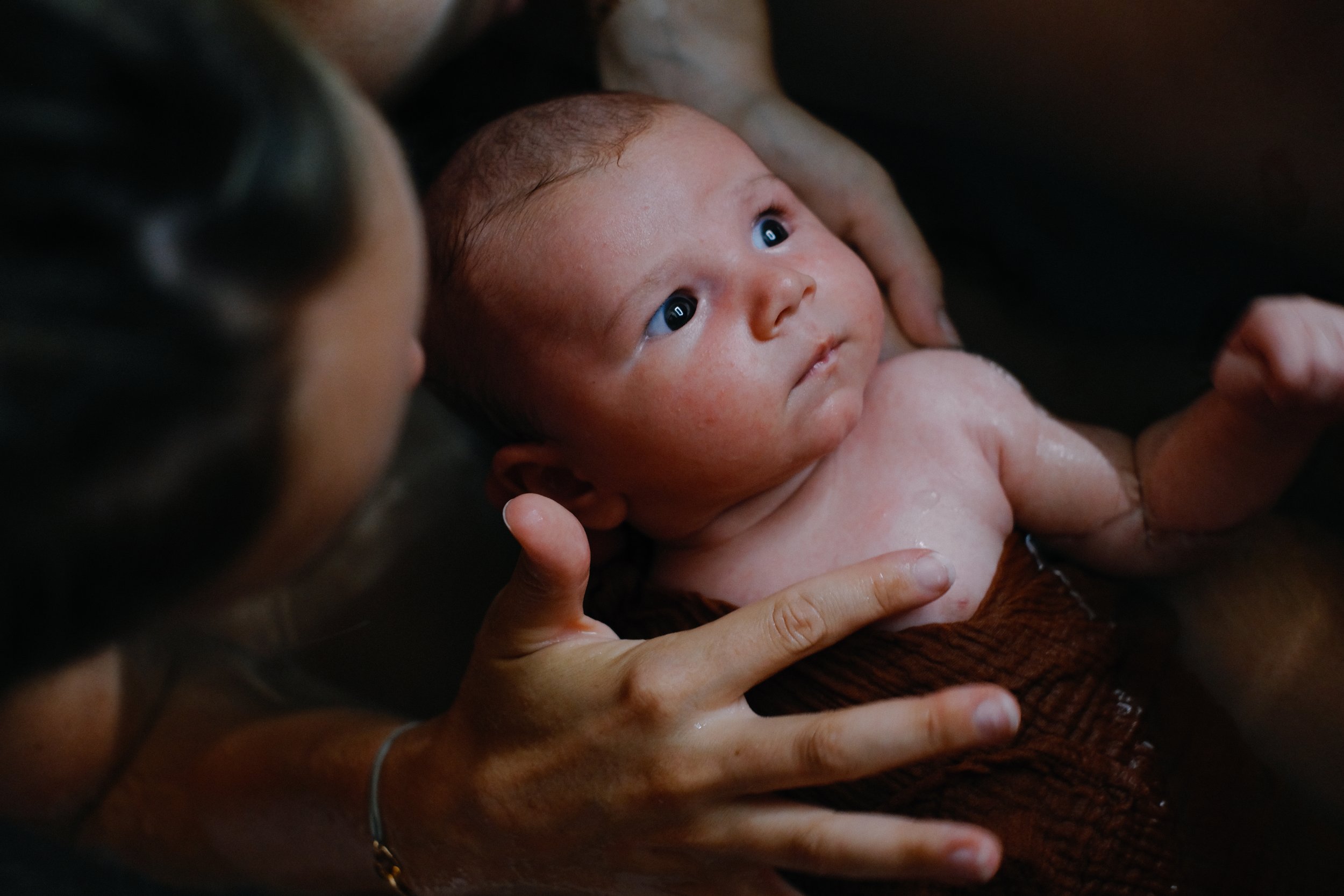 Un bébé regardant vers le haut, tenu dans les bras d'une personne, avec des mains visibles supportant le bébé. La scène est intime et douce.
