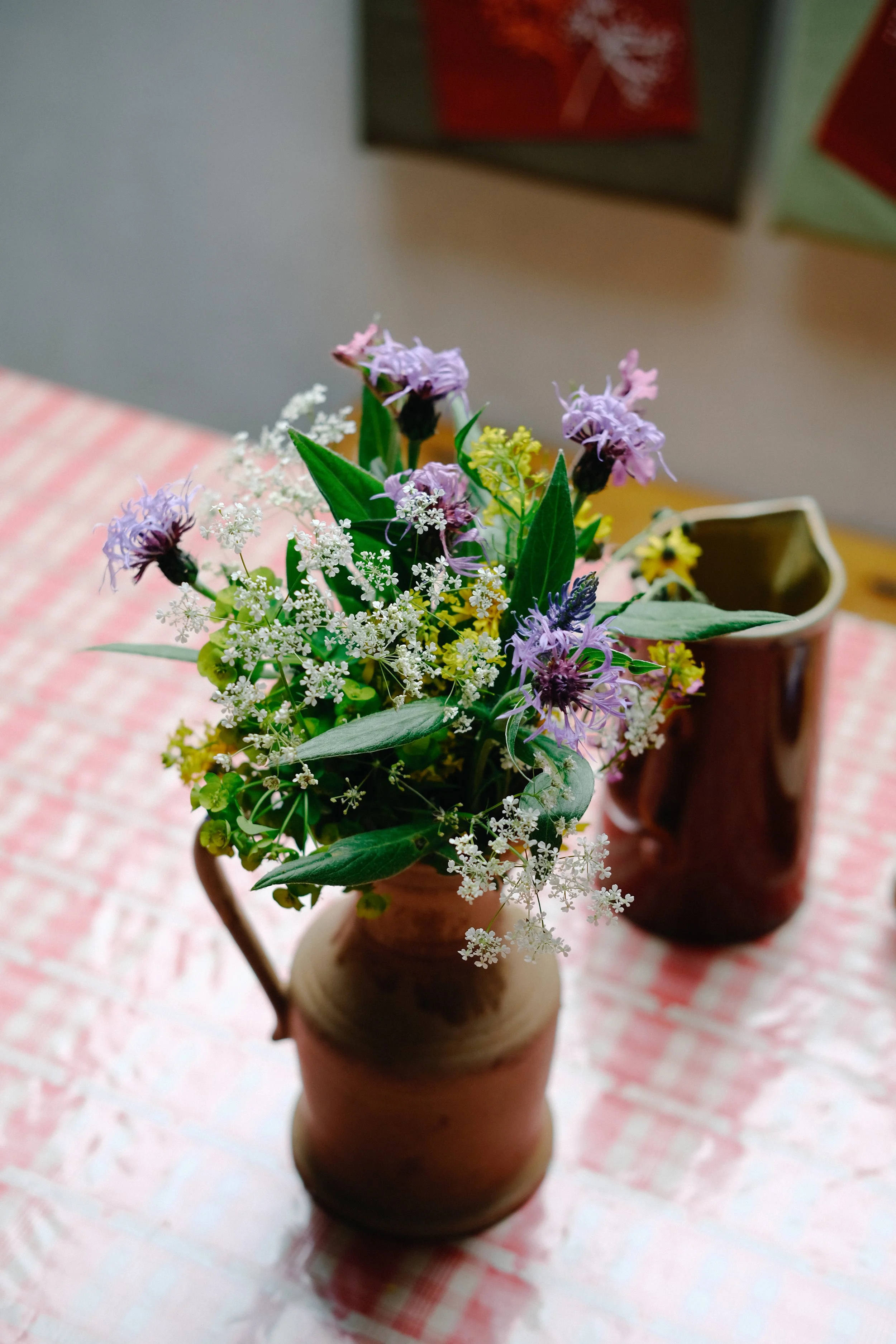 Un bouquet de fleurs colorées dans un vase en terre cuite, posé sur une nappe à carreaux rouges et blancs, avec deux autres vases en arrière-plan.