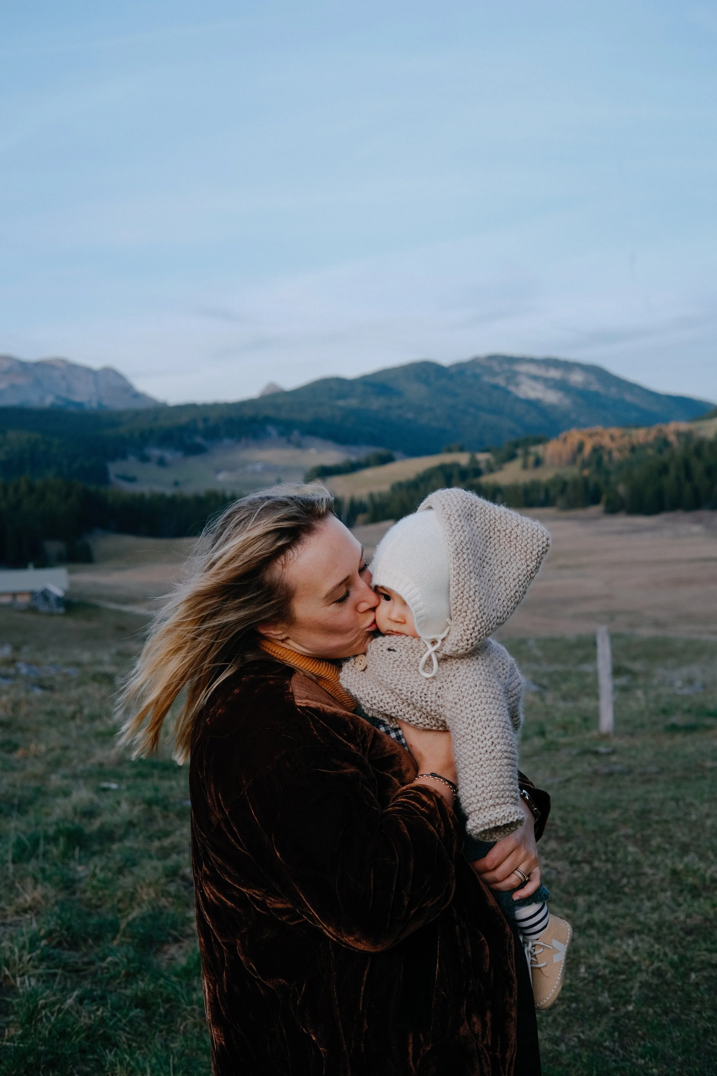 Une femme et un enfant s'embrassent dans un champ avec des montagnes en arrière-plan, ciel clair et paysage naturel.