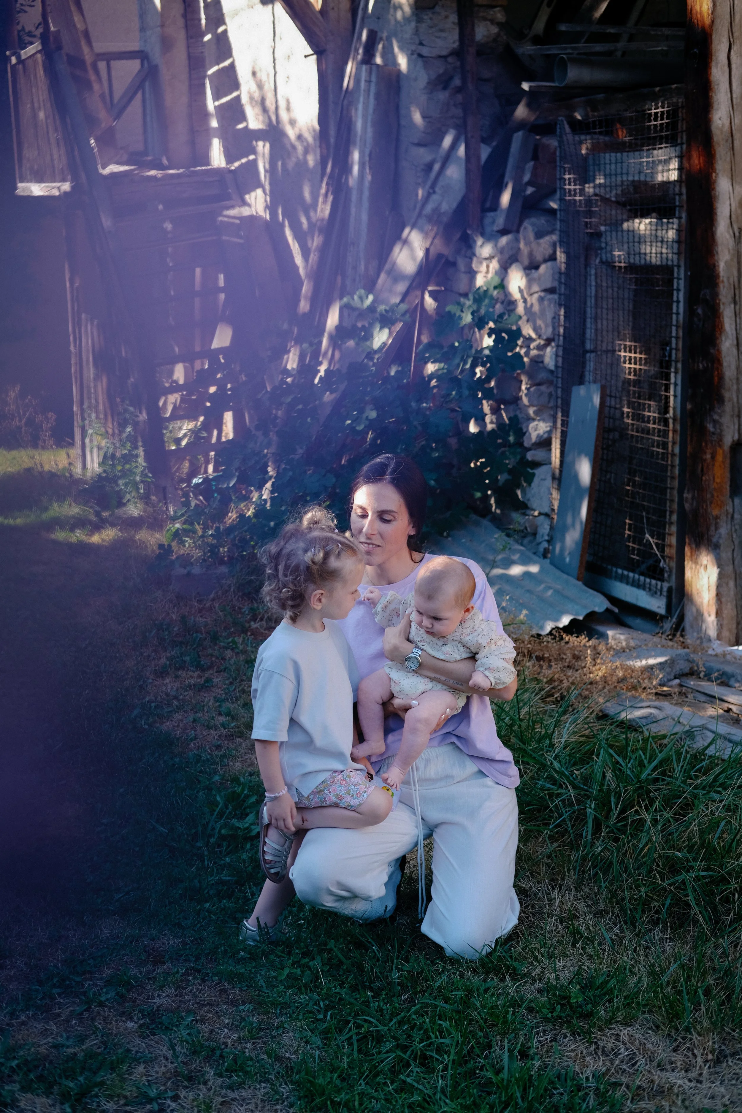 Une femme assise dans un jardin, tenant deux enfants, une fille et un garçon, à l’extérieur, avec des murs en pierre, une clôture métallique et des plantes vertes en arrière-plan.