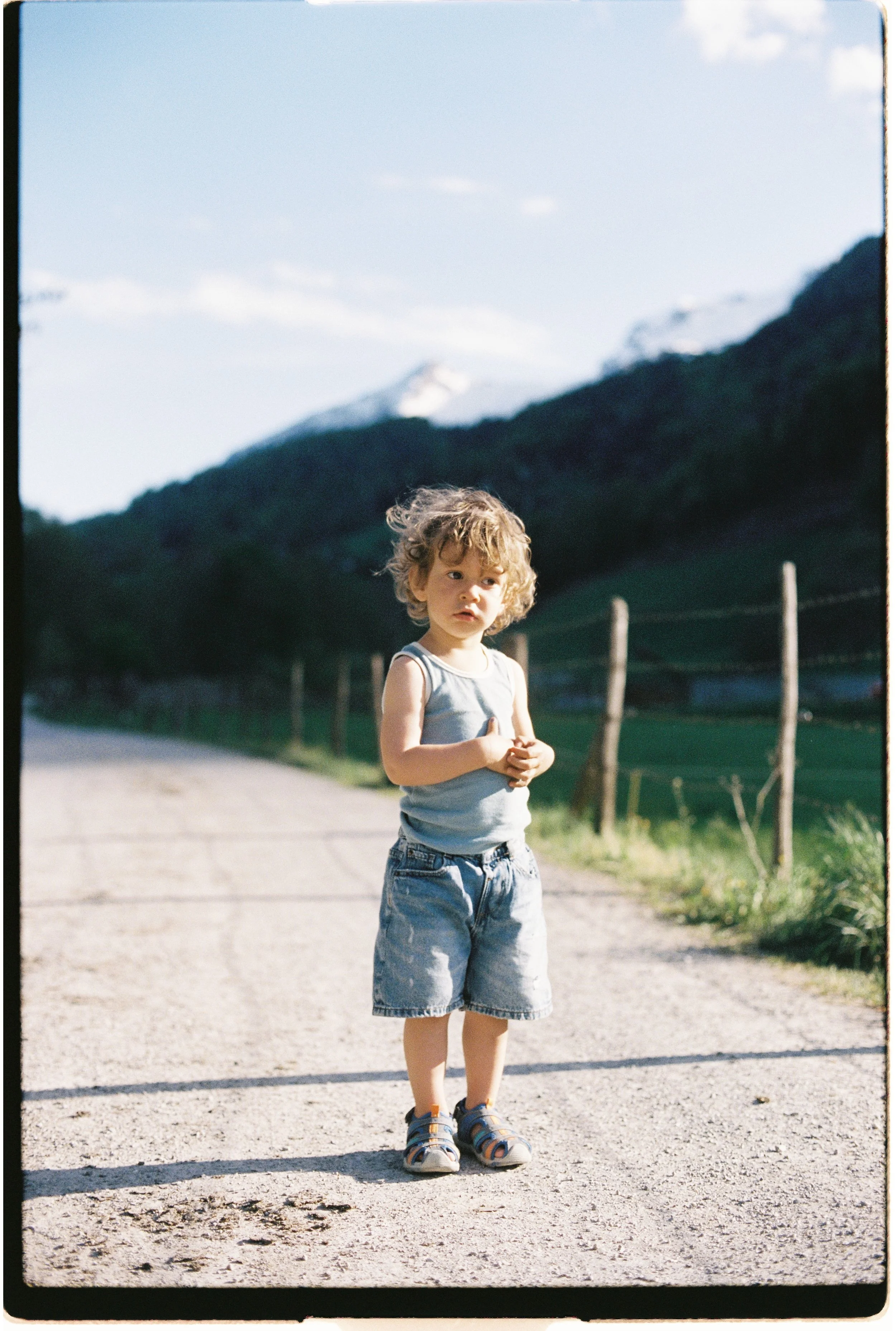 Jeune garçon aux cheveux bouclés portant un t-shirt gris, un short en jean et des sandales, debout sur un chemin en plein air avec un paysage de montagnes en arrière-plan.