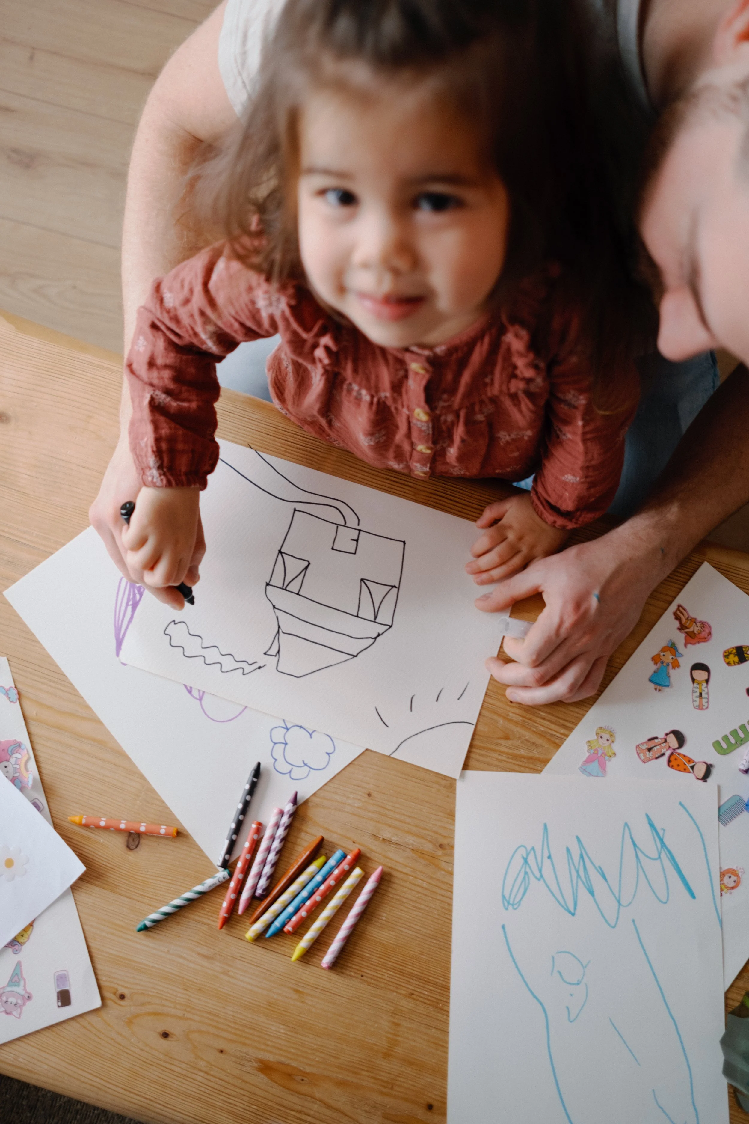 Une jeune fille dessine un bâtiment sur une feuille avec un marqueur noir, entourée de crayons de couleur et de dessins colorés sur la table en bois.