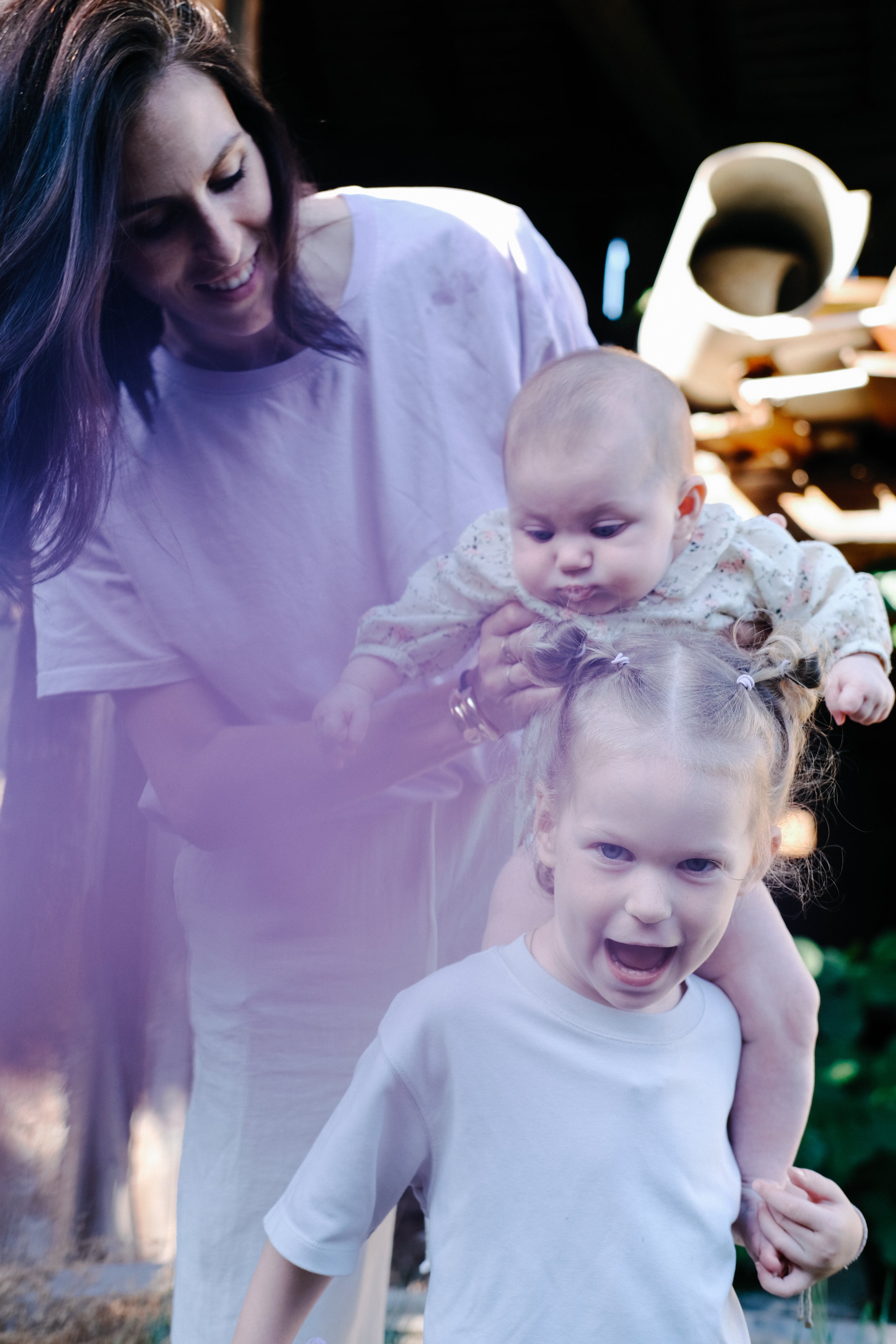 Une femme souriante avec des cheveux violets tenait deux jeunes enfants, un garçon avec une expression joyeuse et une fille regardant un peu perplexe, dans un environnement extérieur avec des arbres en arrière-plan.