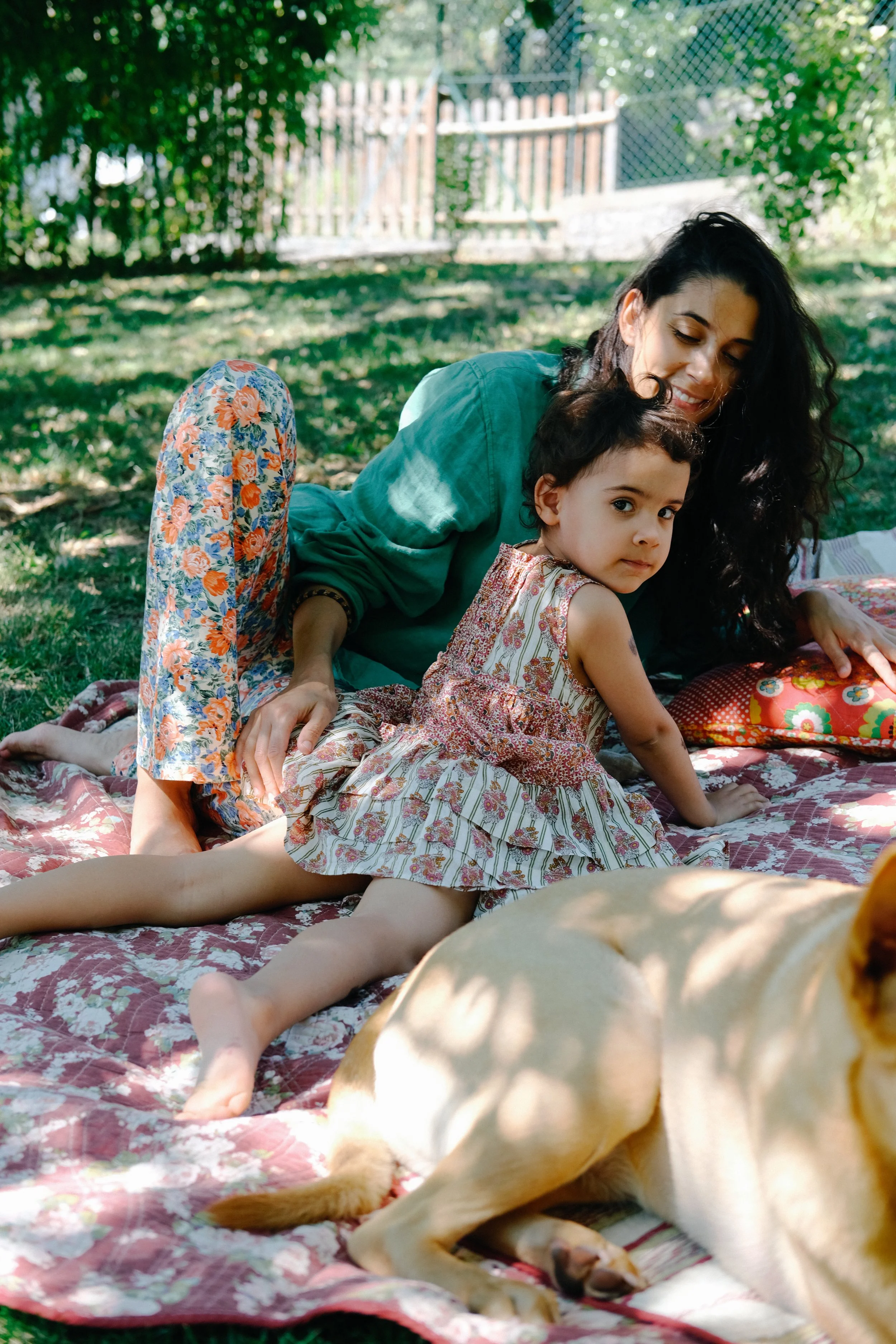 Une femme et un enfant jouent avec un chien dans un jardin ensoleillé, allongés sur une couverture à motifs floraux.
