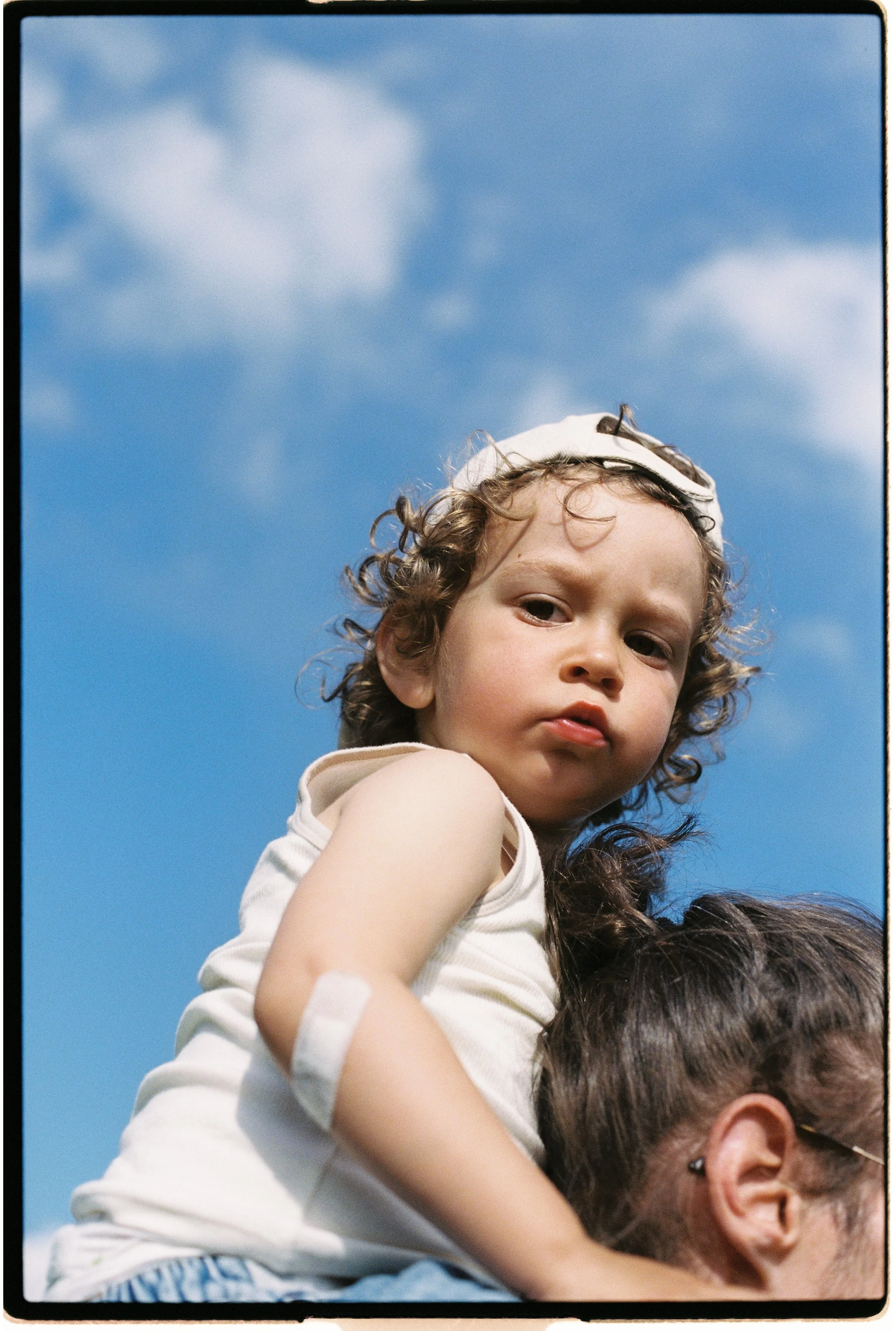 Un enfant aux cheveux bouclés, portant un chapeau blanc, est assis sur une personne adulte, avec un ciel bleu en arrière-plan.