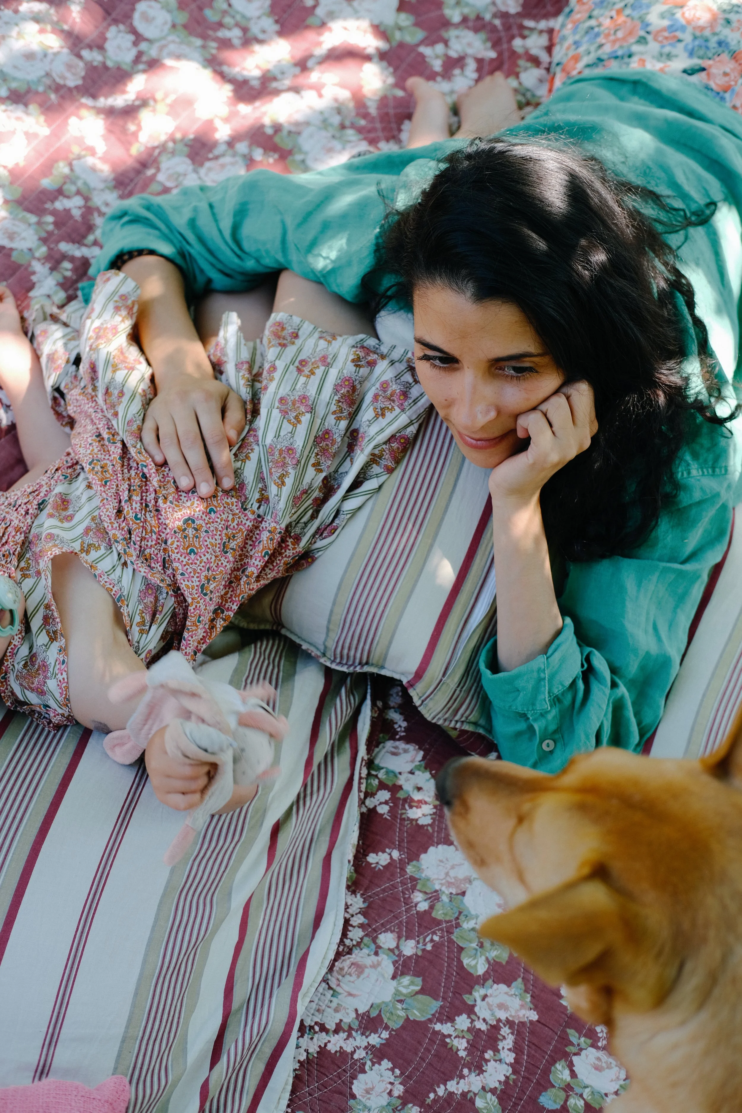 Une femme et une enfant allongées sur une couverture à motifs floraux, regardant un chien proche d'elles, dans un cadre extérieur ensoleillé.