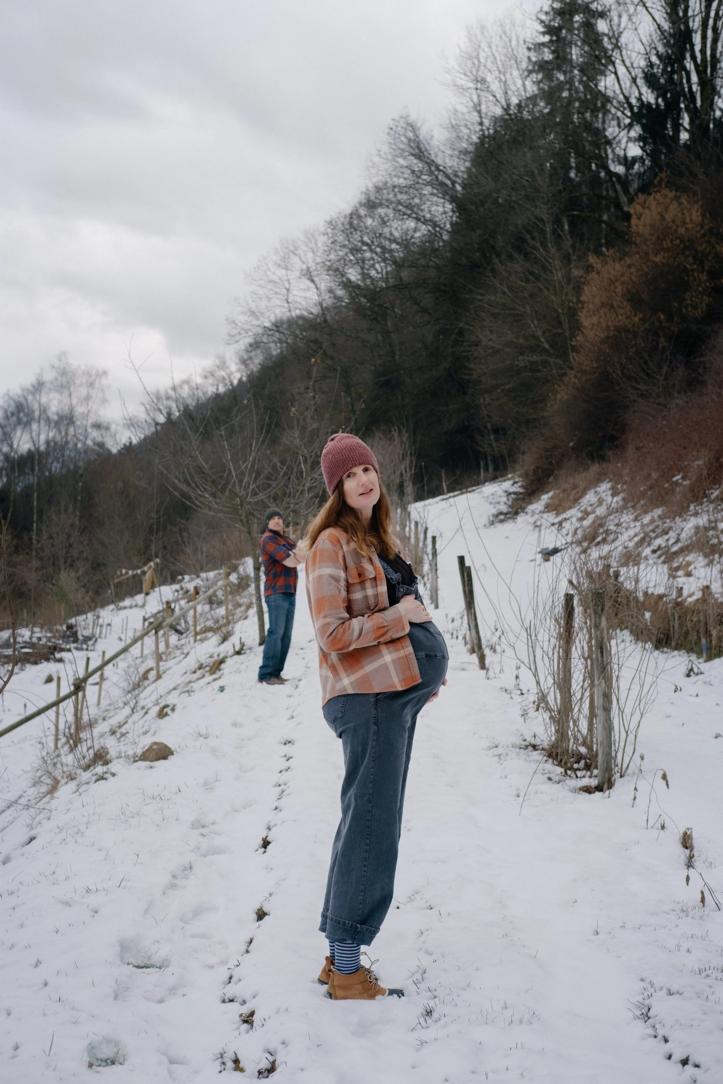 Une femme enceinte portant un chapeau en laine marron, un manteau à carreaux, des jeans et des bottines, se tient dans la neige avec un paysage boisé en arrière-plan. Un homme se tient derrière elle dans la neige, portant une veste à carreaux et un b