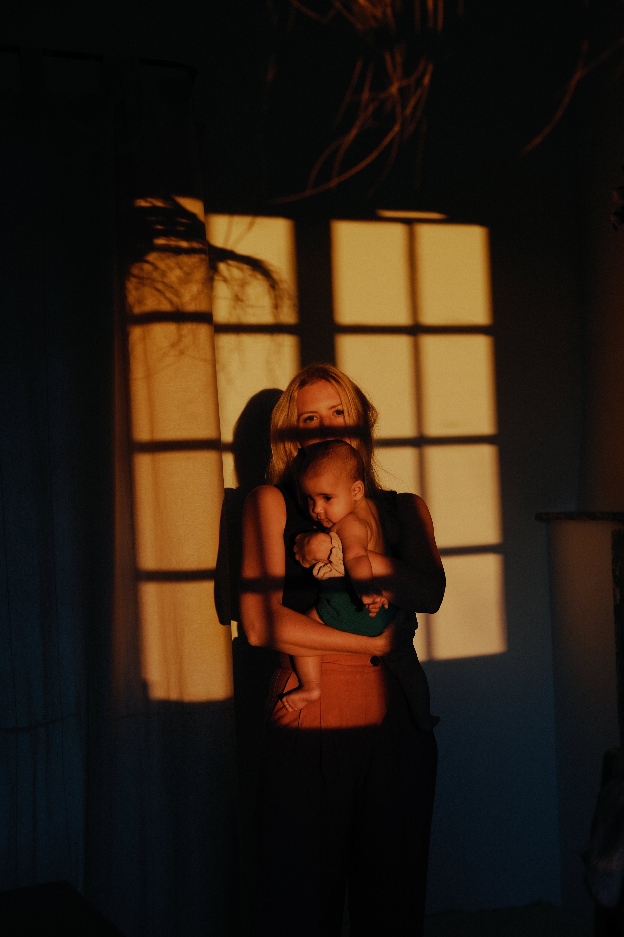 Une femme blonde tient un bébé dans ses bras, la lumière du soleil filtre à travers une fenêtre, jetant des ombres sur le mur. L'ambiance est chaleureuse et intime.