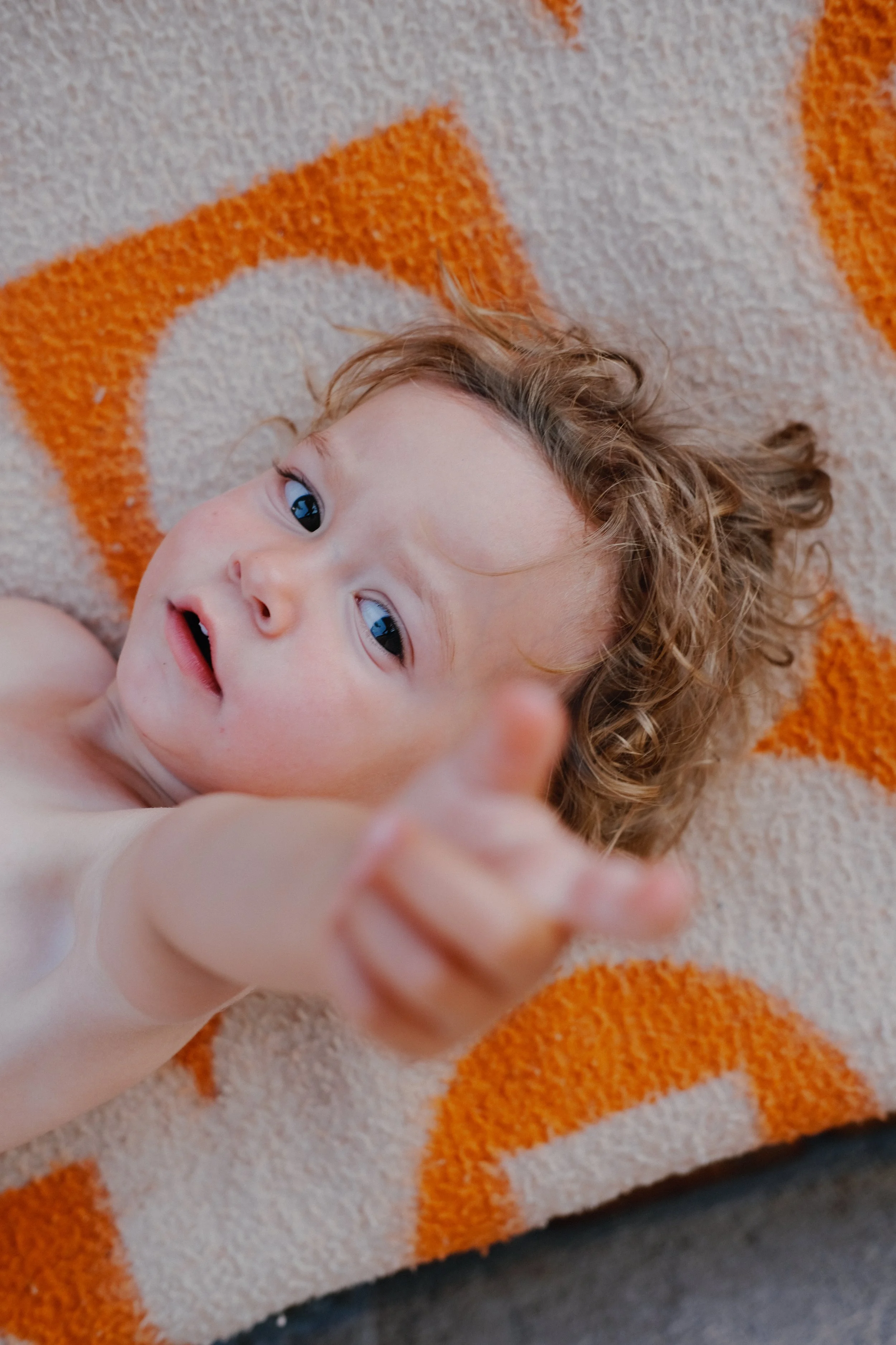 Un jeune enfant aux cheveux bouclés et yeux bleus allongé sur un tapis coloré, regardant vers la caméra avec un sourire, bébé de profil avec un bras tendu