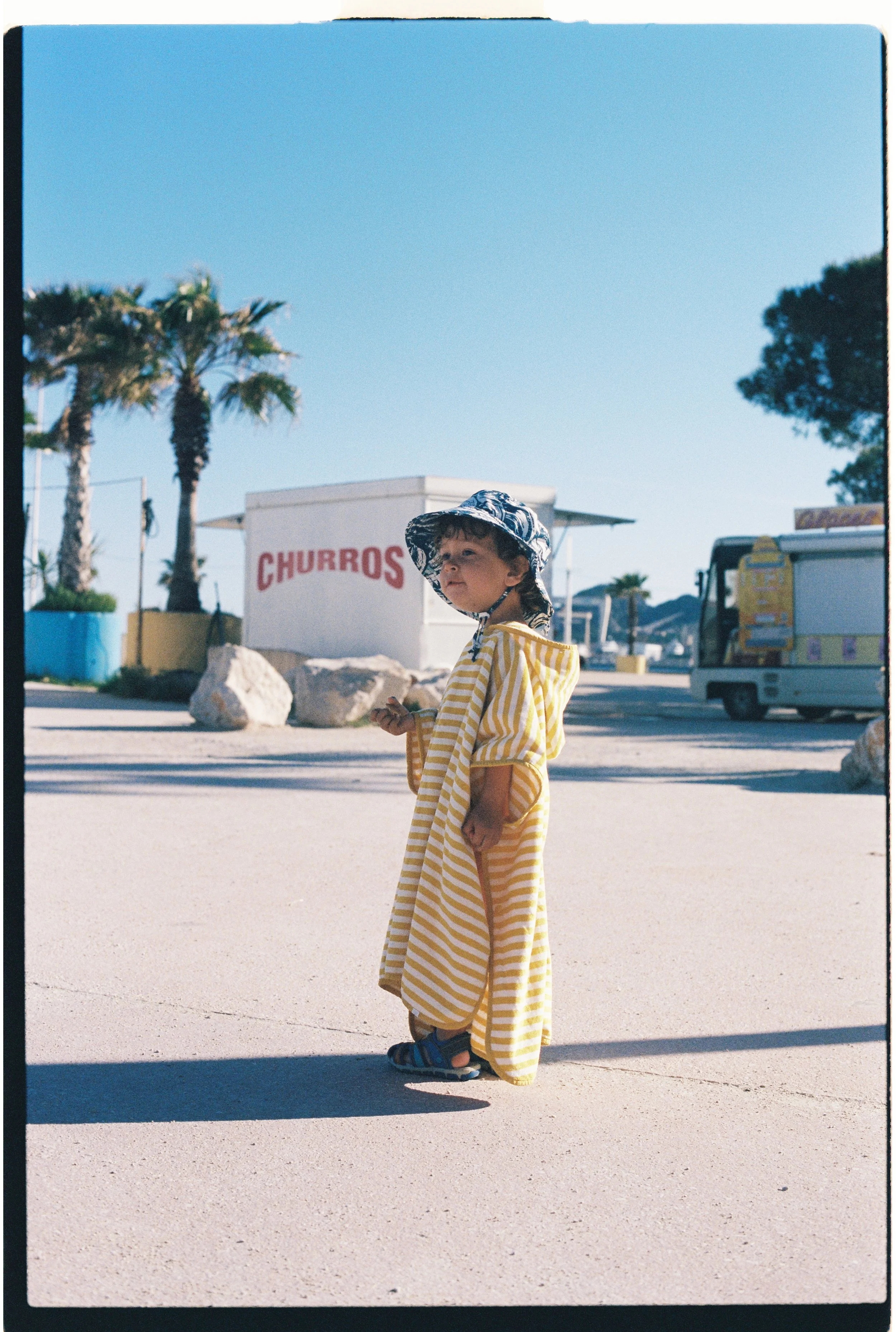 Un enfant portant une tunique rayée jaune et blanc, un chapeau assorti, et des sandales, se tient sur un sol en béton clair, avec un stand de churros et des arbres en arrière-plan dans un environnement en plein air.