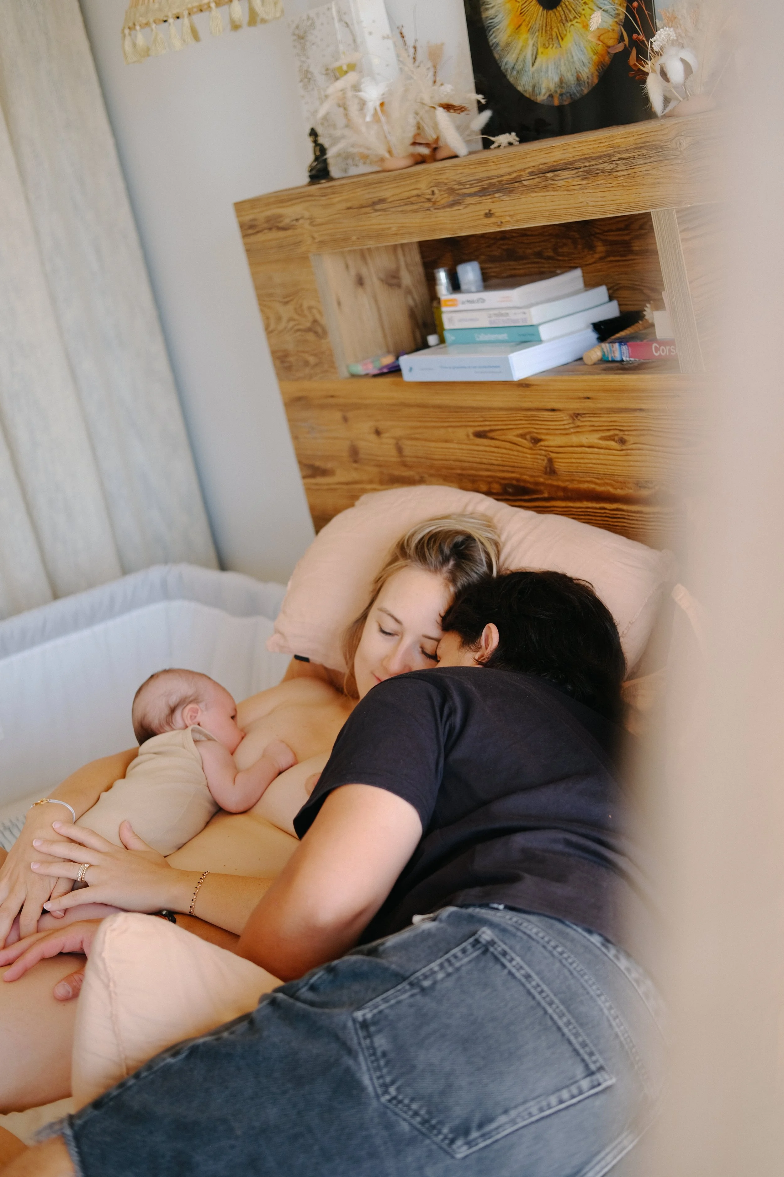 Une femme nue en train d'allaiter un bébé dans un lit, accompagnée d'un homme qui se repose à côté, dans une chambre chaleureuse avec un mur en bois et des livres