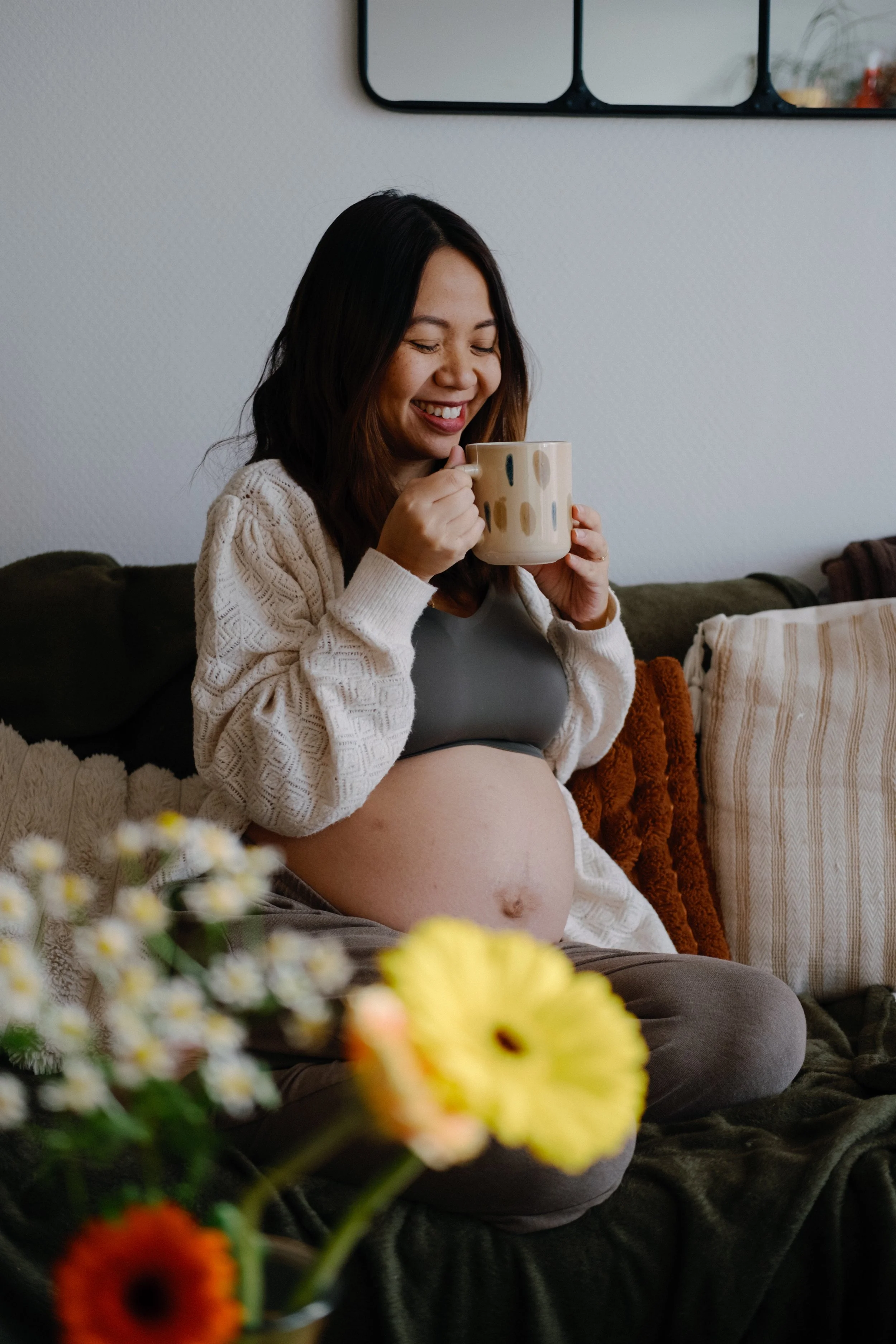 Une femme enceinte assise sur un canapé, tenant une tasse, souriant avec des yeux fermés, dans un intérieur décoré avec des coussins et des fleurs en premier plan.