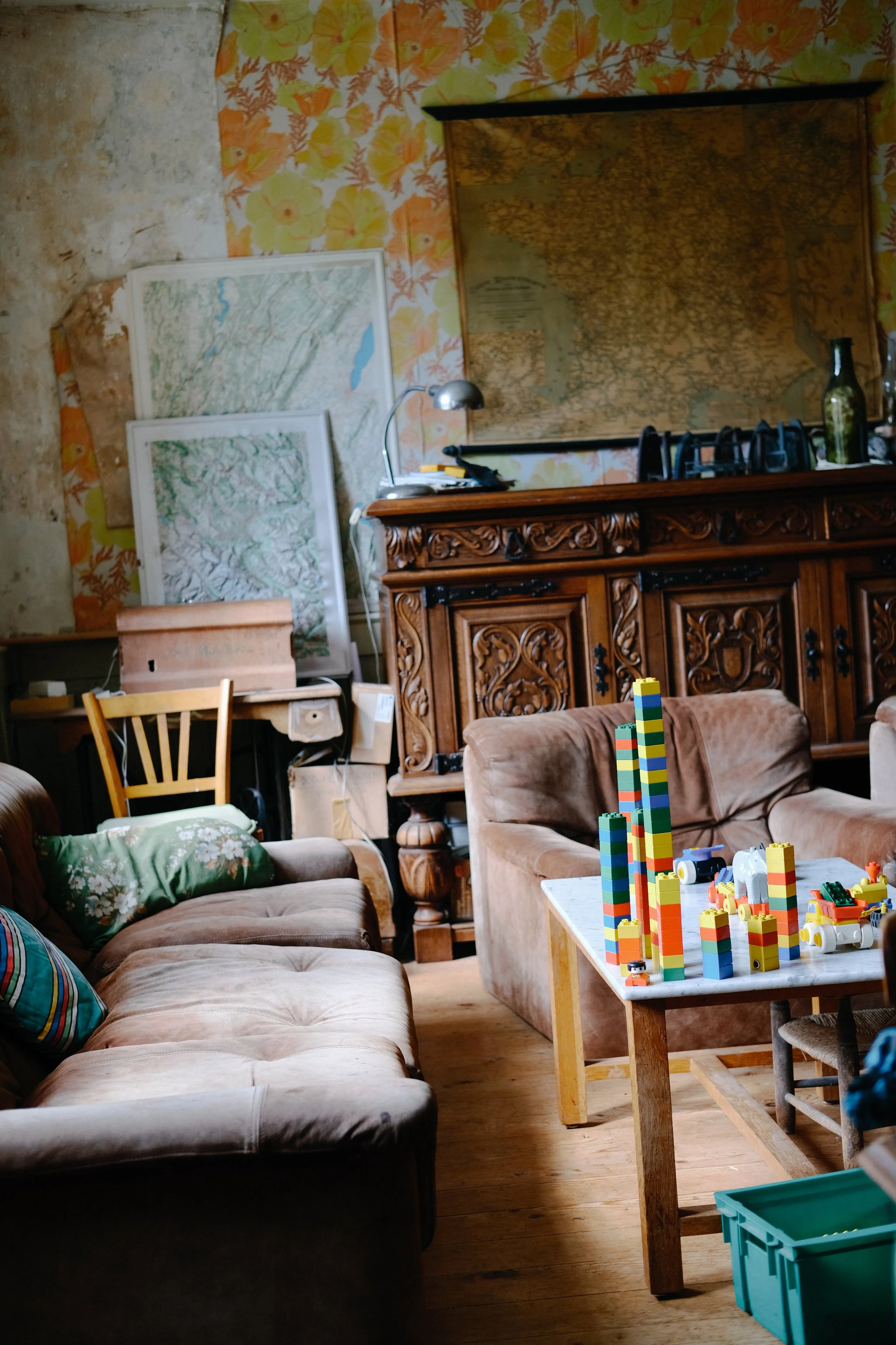 Un salon avec des canapés, une table avec des blocs de construction colorés, une étagère en bois sculptée, des cartes ou planches accrochées au mur, et des objets divers comme une bouteille et une lampe de bureau.