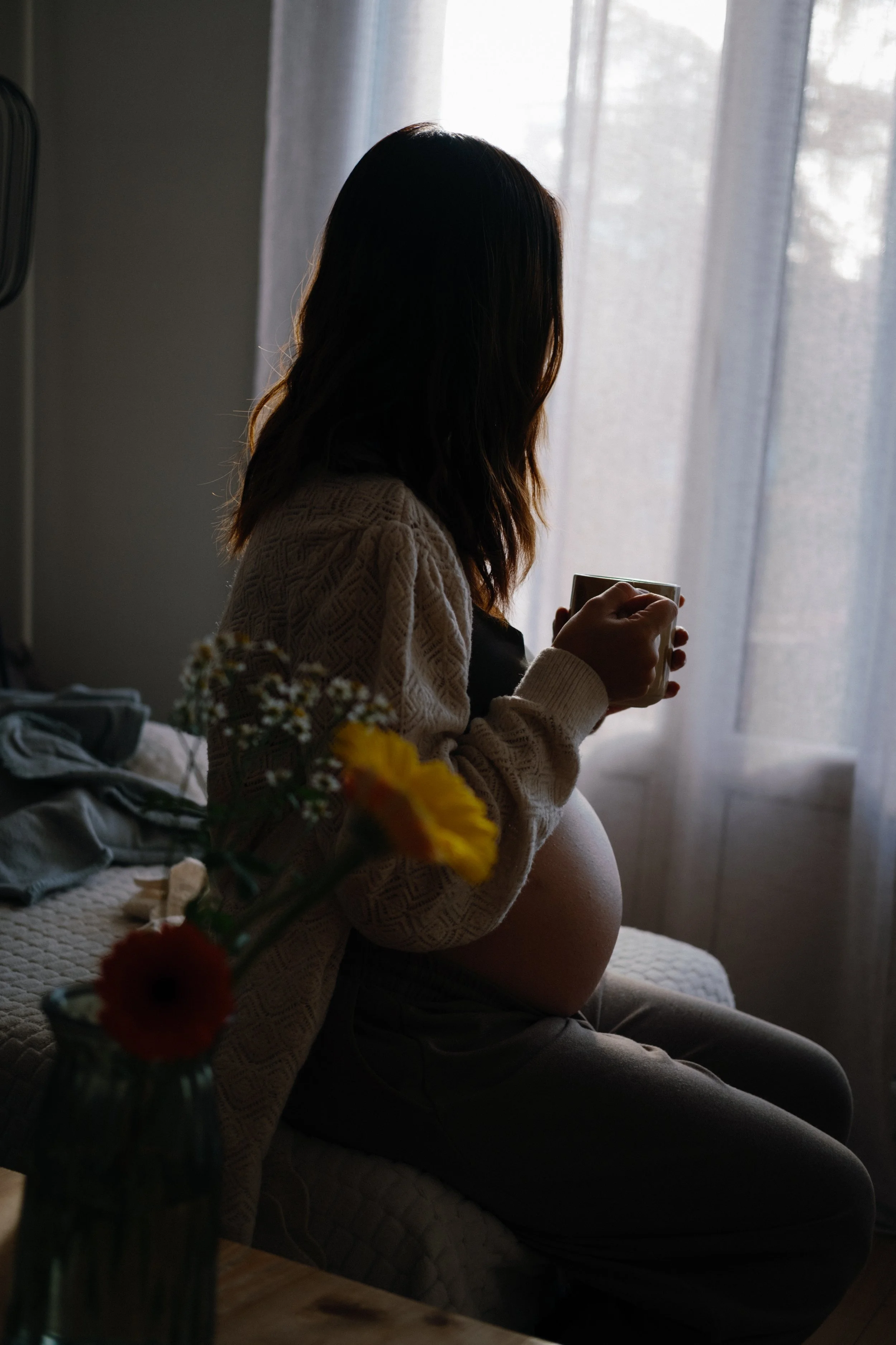 Une femme enceinte assise sur un lit, tenant une tasse, dans une pièce lumineuse avec des rideaux translucides.