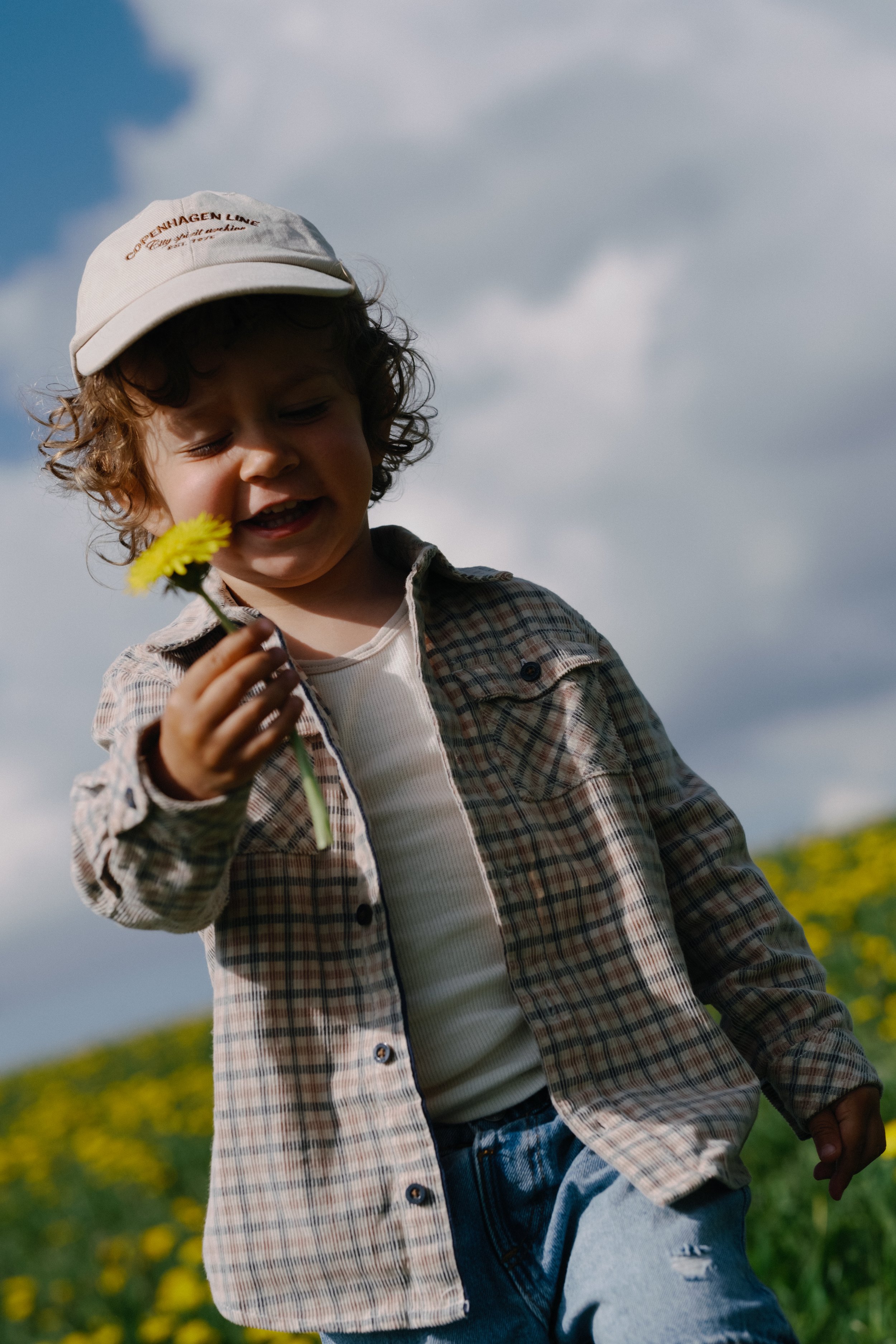 Un jeune garçon avec des cheveux bouclés, portant une casquette blanche, un manteau à carreaux, un t-shirt blanc et un jean, qui tient une fleur jaune, dans un champ avec un ciel nuageux.