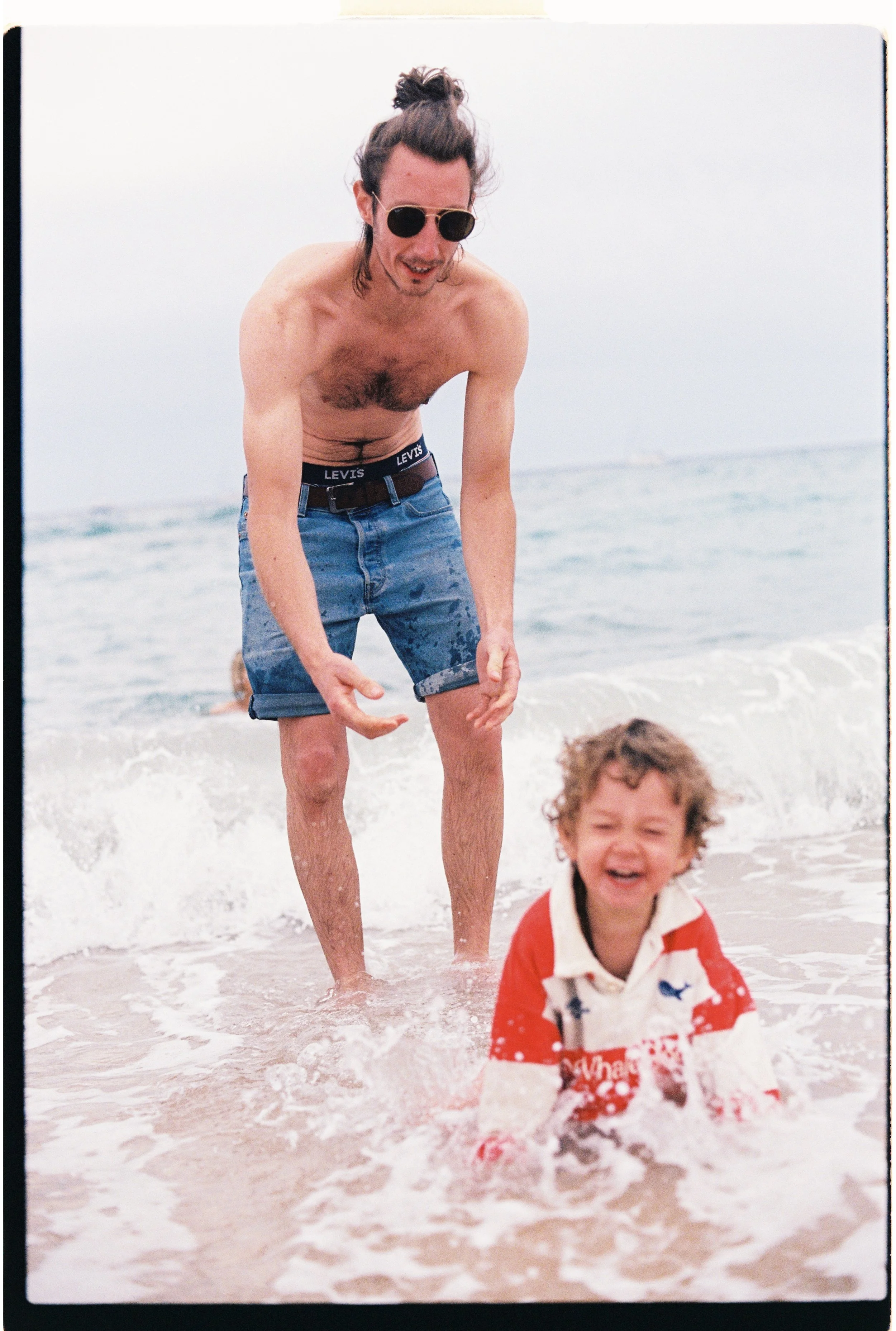 Un homme avec des lunettes de soleil, torse nu, en short en jean, joue dans la mer avec deux enfants à la plage, une enfant en chemise à rayures rouges et blanches, l'autre personne floue en arrière-plan.