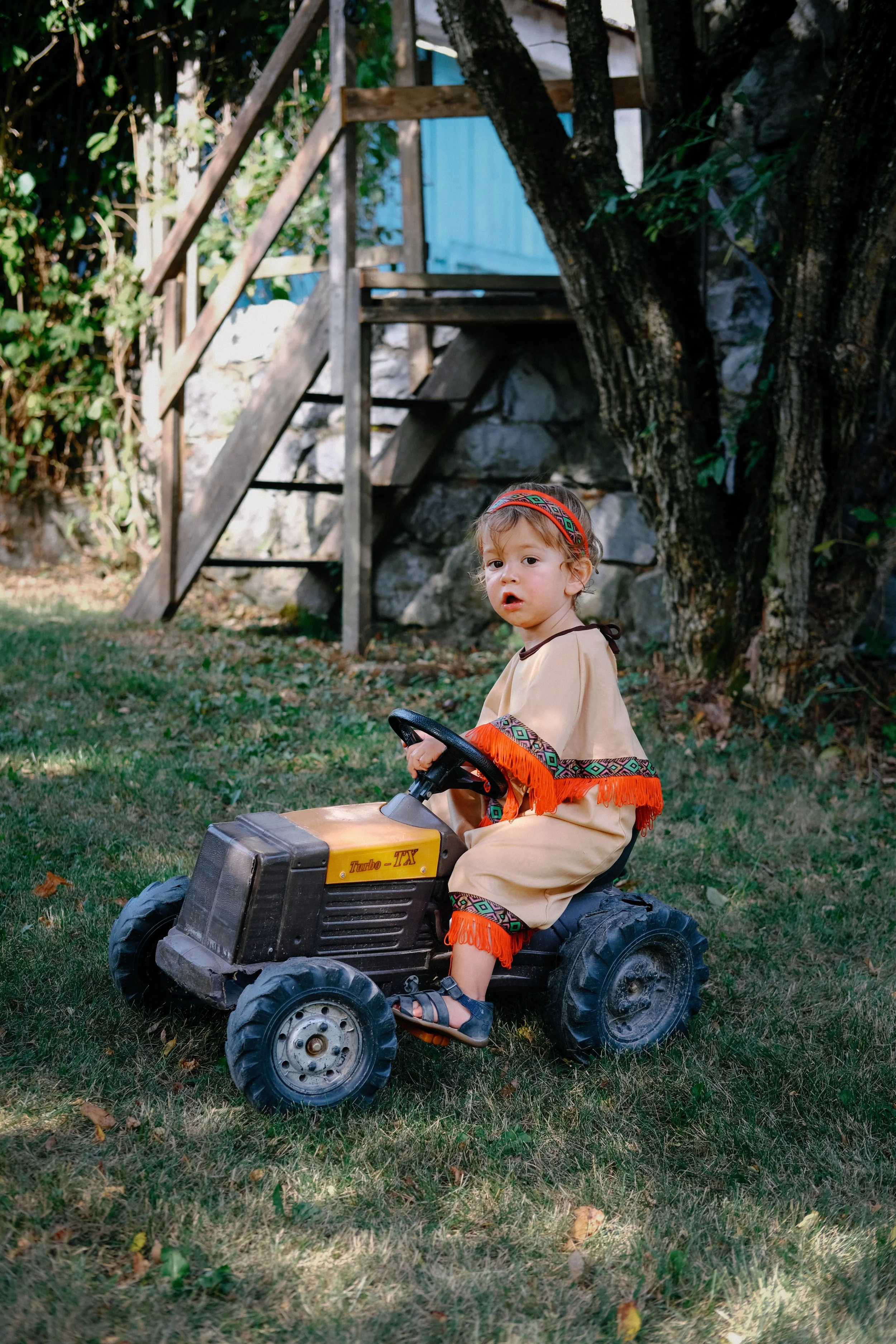 Un jeune enfant assis sur une petite voiture électrique dans un jardin avec des arbres et un escalier en bois en arrière-plan, portant un costume traditionnel avec des motifs colorés.