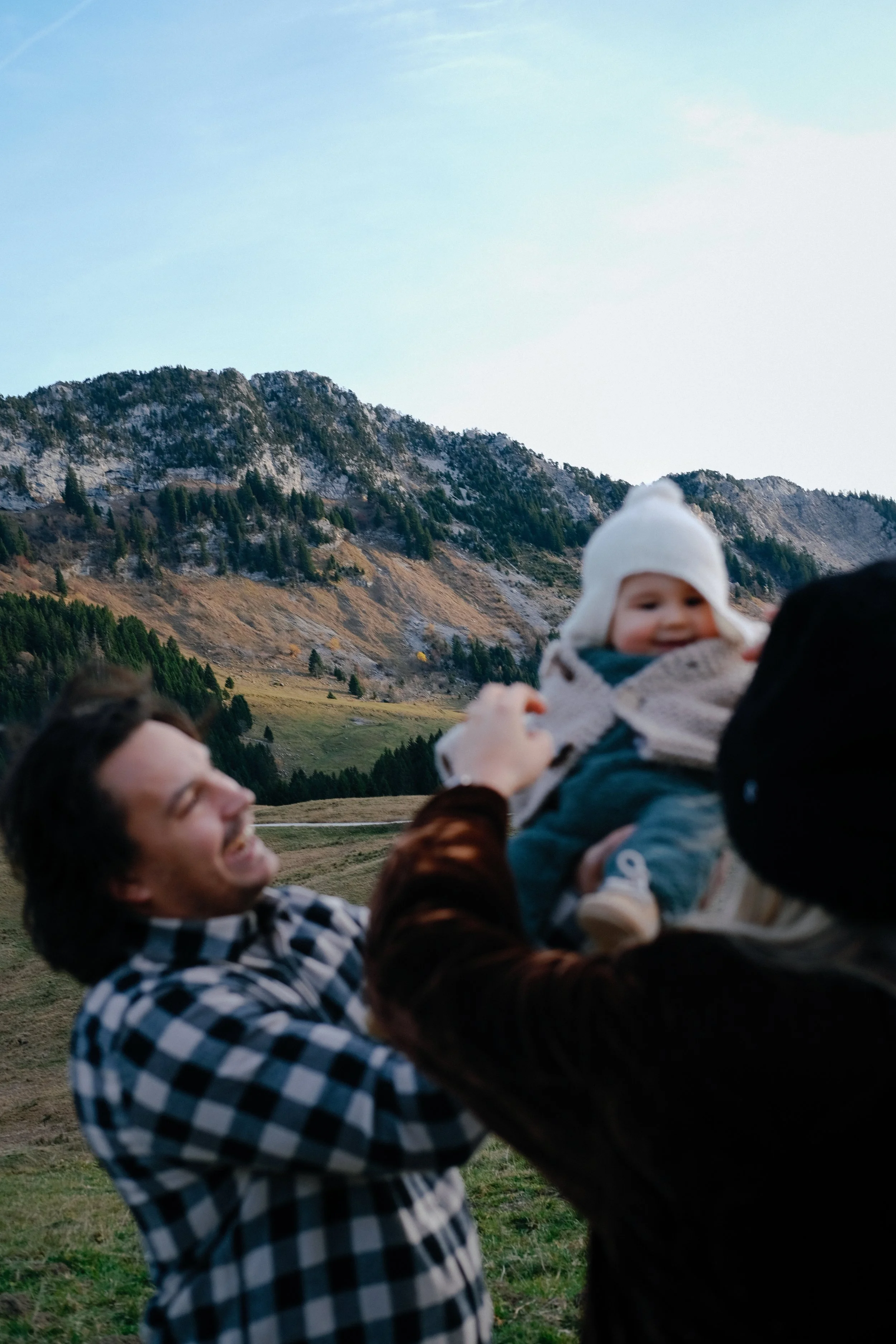 Une personne souriante tient un bébé dans ses bras, avec un paysage de montagnes et de forêt en arrière-plan.