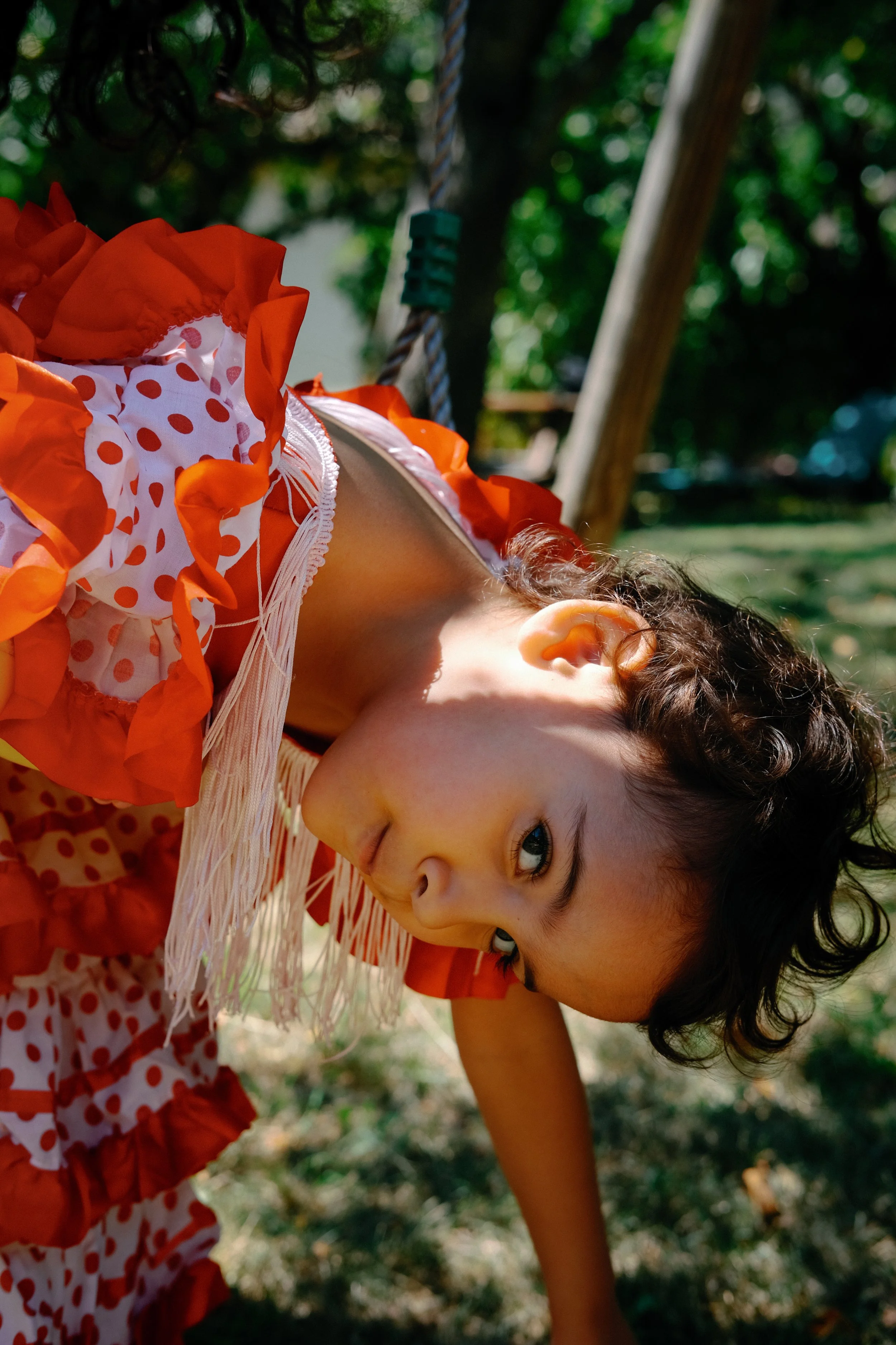 Un enfant avec des cheveux bouclés, portant un costume de flamenco coloré, est allongé par terre dans un parc ombragé, regardant vers l'objectif.