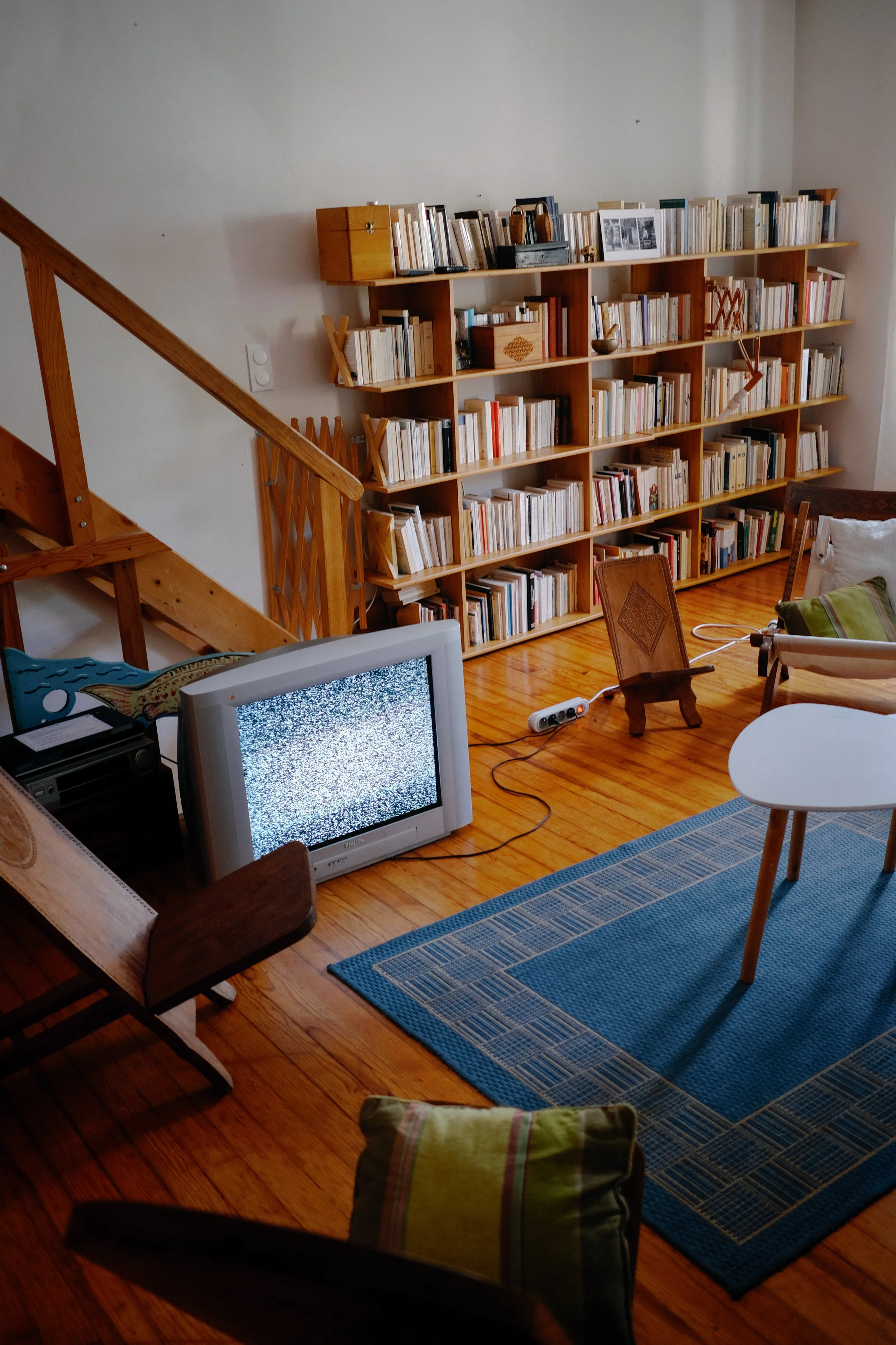 Une pièce avec bibliothèque en bois, tapis bleu, fauteuil, télévision, et plusieurs coussins.