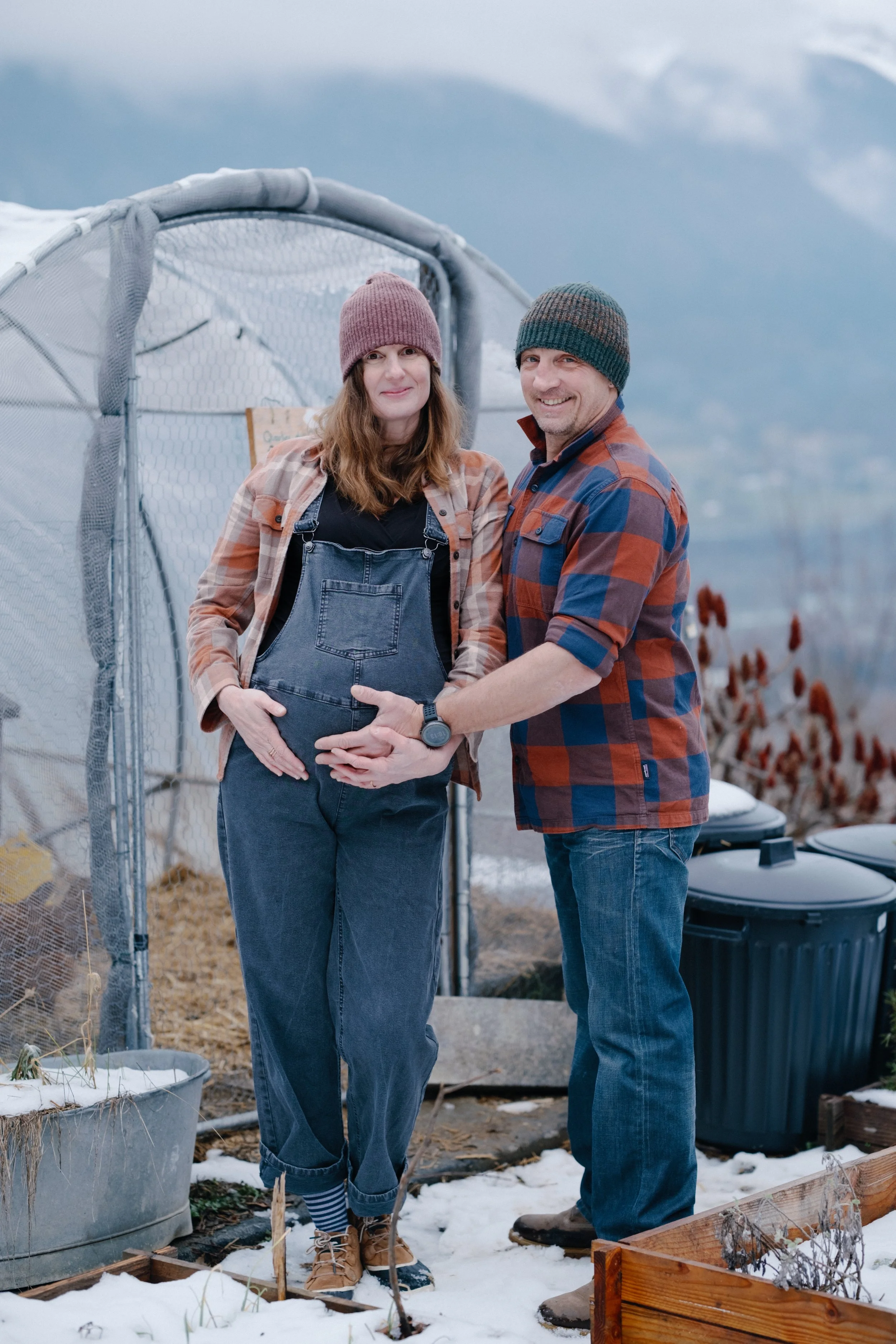 Un couple, un homme et une femme, posent dans un jardin en hiver, la femme est enceinte, entourés de potagers et d'outils de jardin, avec des montagnes enneigées en arrière-plan.