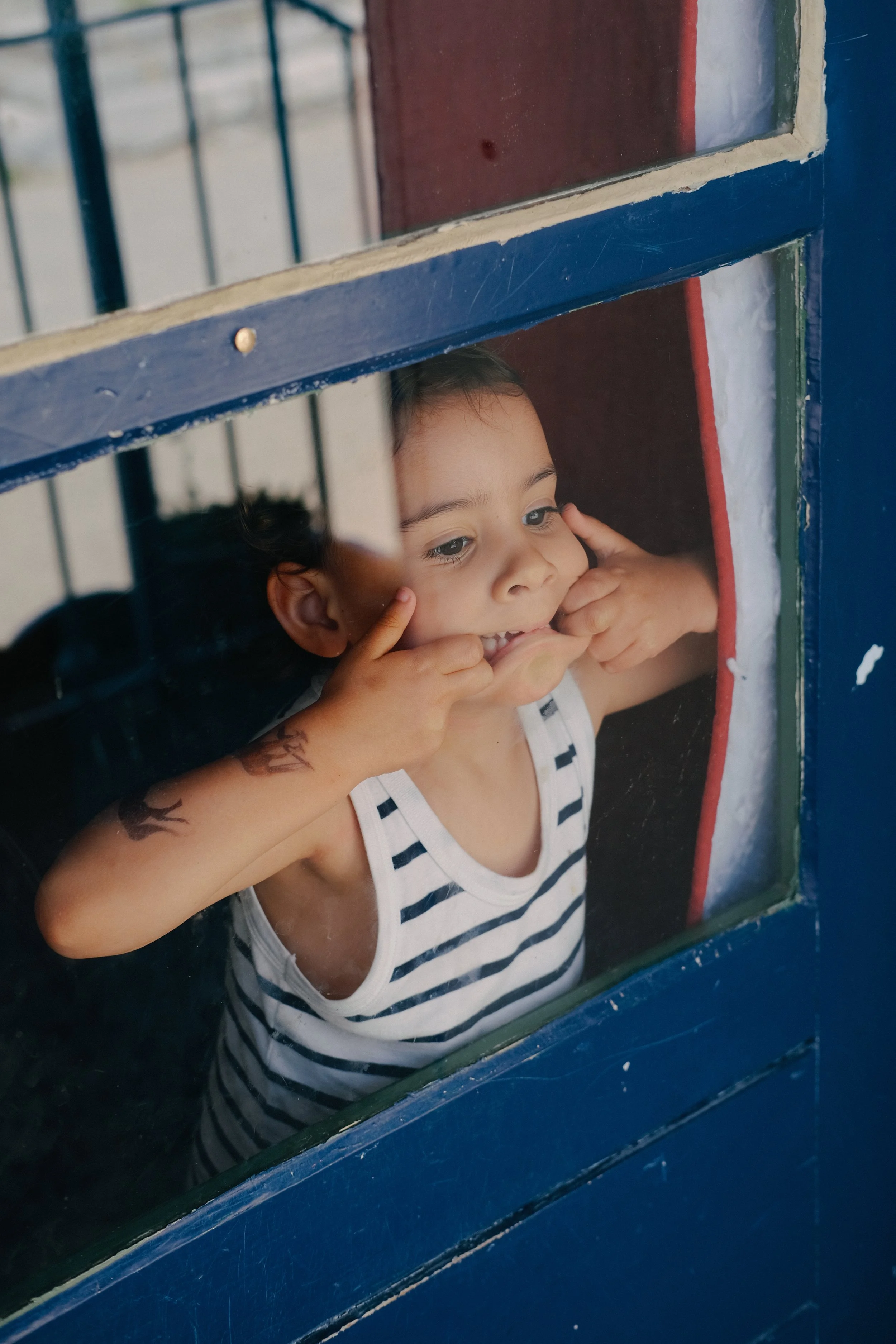 Jeune garçon regardant par une fenêtre en bois peint en bleu, portant un t-shirt à rayures et ayant des tatouages sur les bras.