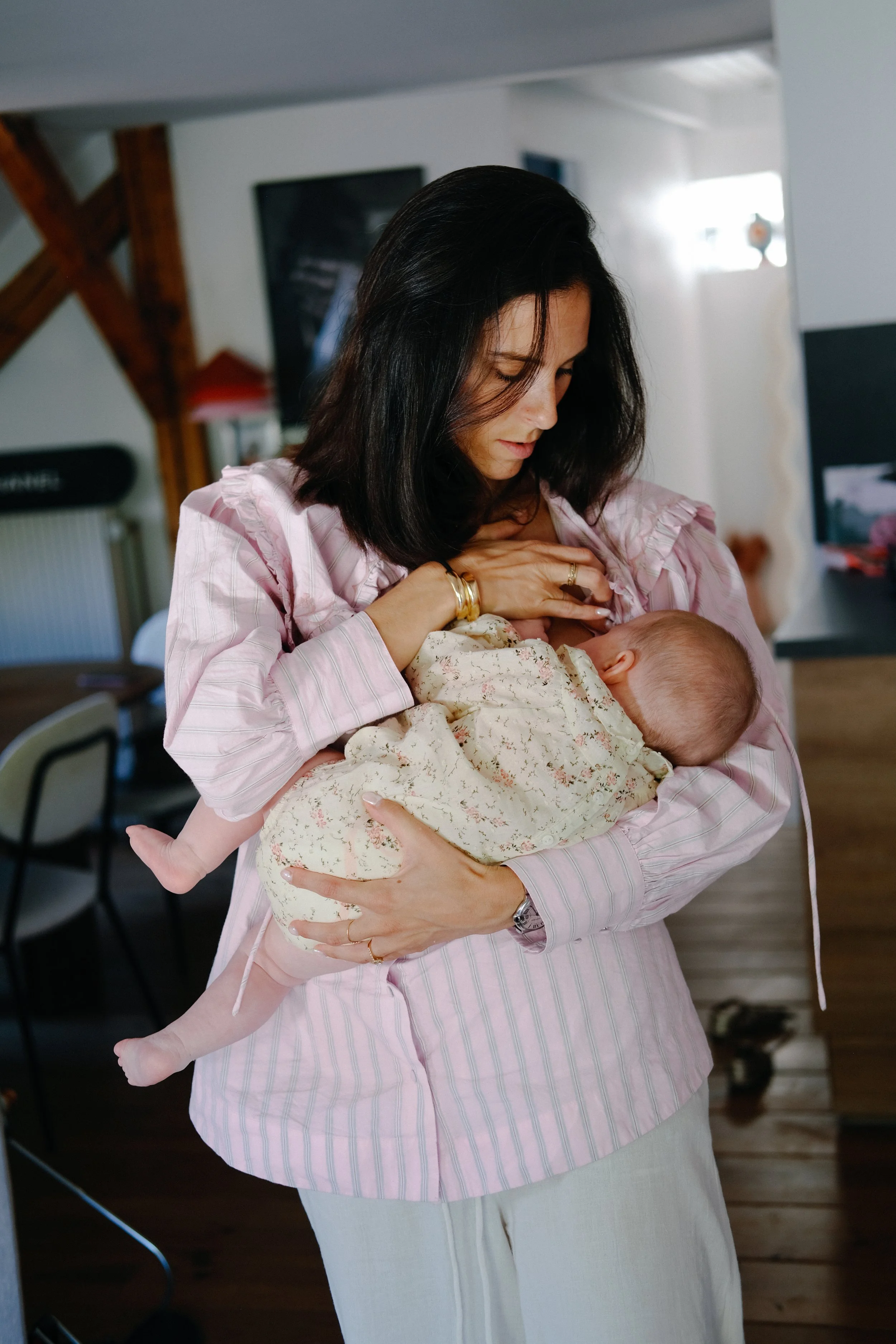 Une femme avec des cheveux noirs courts, portant une chemise rose rayée, tient un bébé dans ses bras, dans une pièce intérieure.