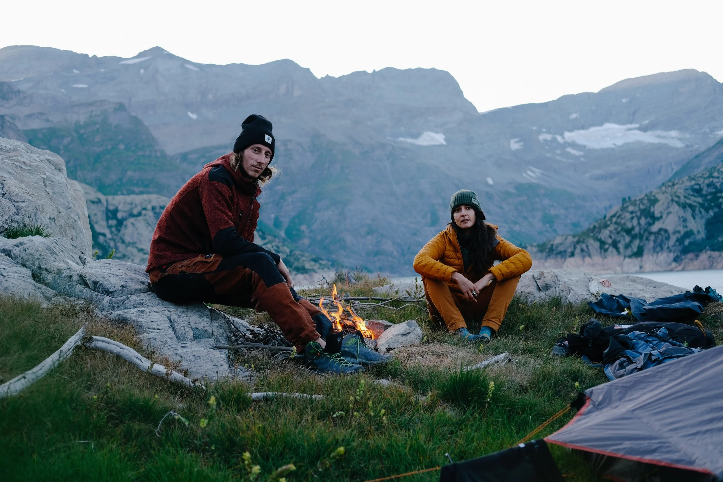 Deux personnes assises près d'un feu de camp dans un paysage montagneux, un homme portant un bonnet noir et une veste rouge, une femme portant un bonnet vert et une veste jaune, avec des montagnes en arrière-plan.