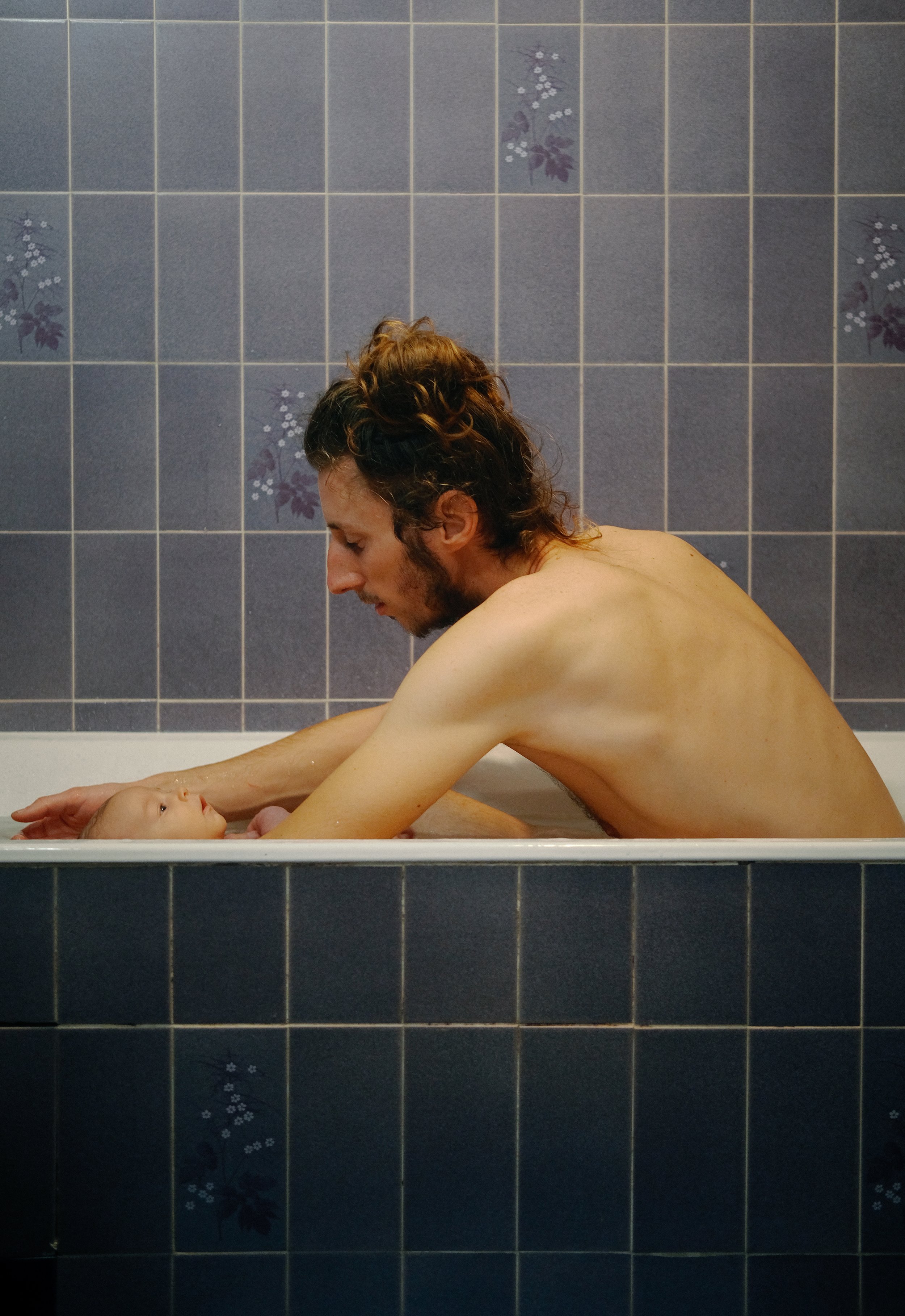 Un homme avec des cheveux bouclés colorés en blond et brun est dans une baignoire, regardant un bébé allongé dans l'eau, avec un mur en carreaux bleus en arrière-plan.