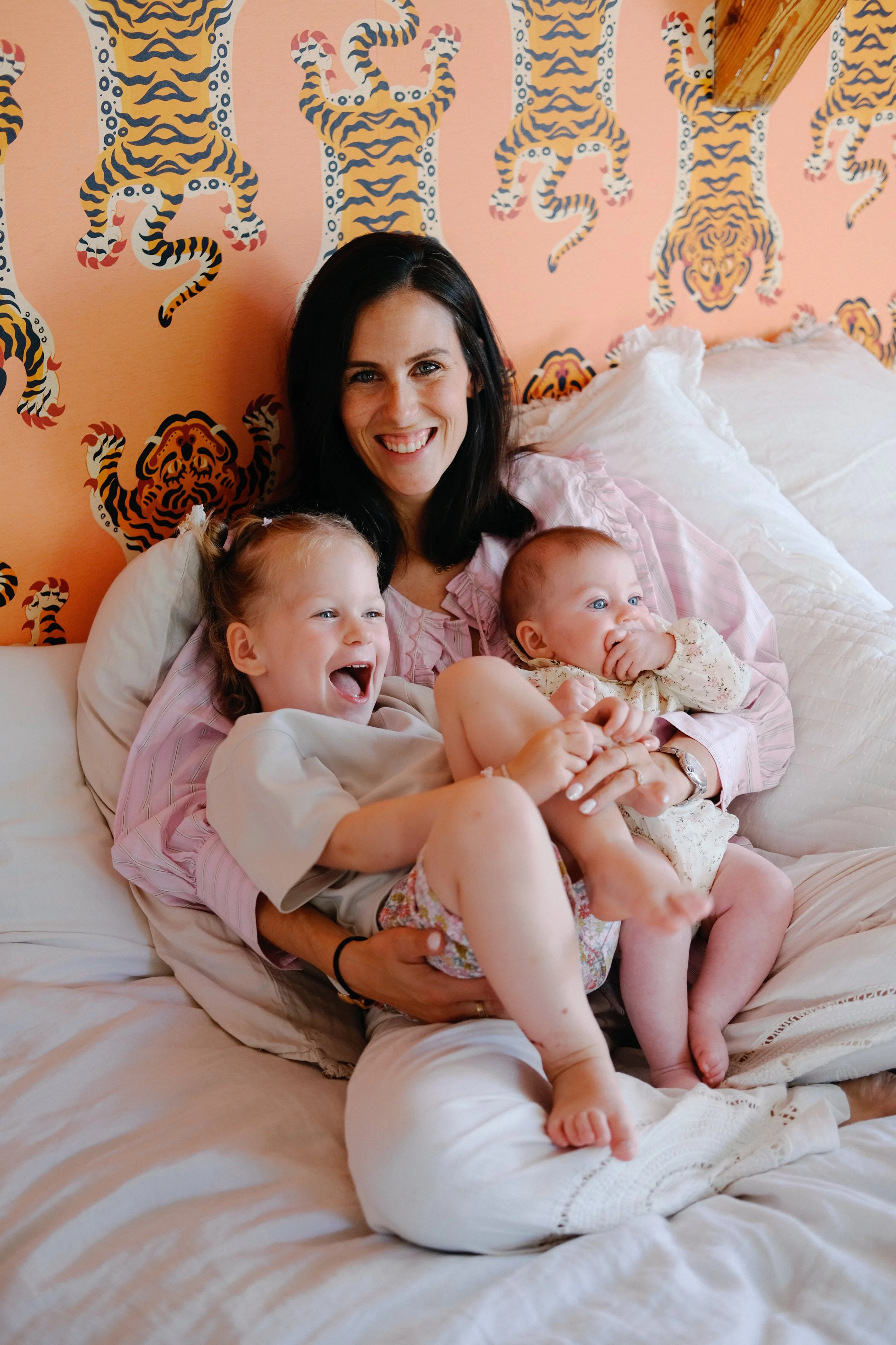 Une femme avec deux jeunes enfants, tous souriants, assis sur un lit avec des oreillers, devant un mur avec un motif de tigres stylisés en noir, blanc, gris et orange sur un fond beige