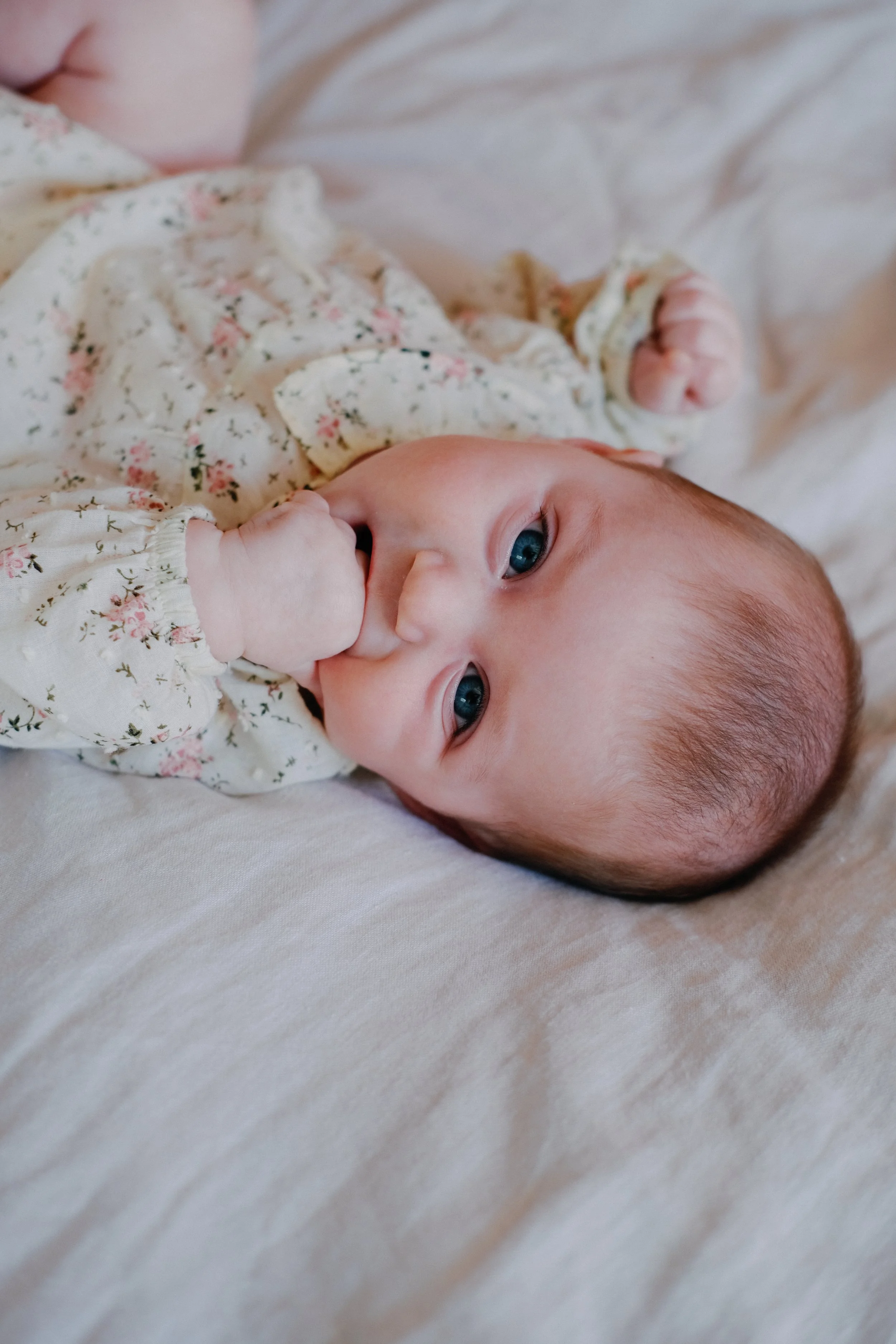 Un bébé allongé sur un lit, portant un pyjama à fleurs, qui a les yeux bleus et qu'il est en train de sucer son pouce.
