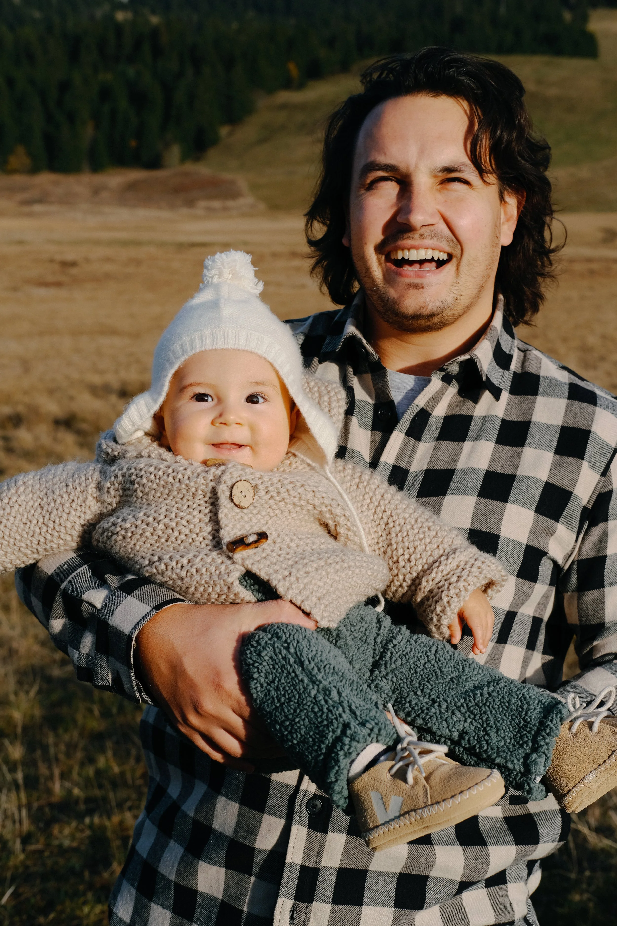 Un homme tient un bébé dans ses bras, tous deux souriants. L'homme porte une chemise à carreaux, le bébé porte un bonnet blanc, un pull beige à boutons et des pantalons gris avec des chaussures beige.