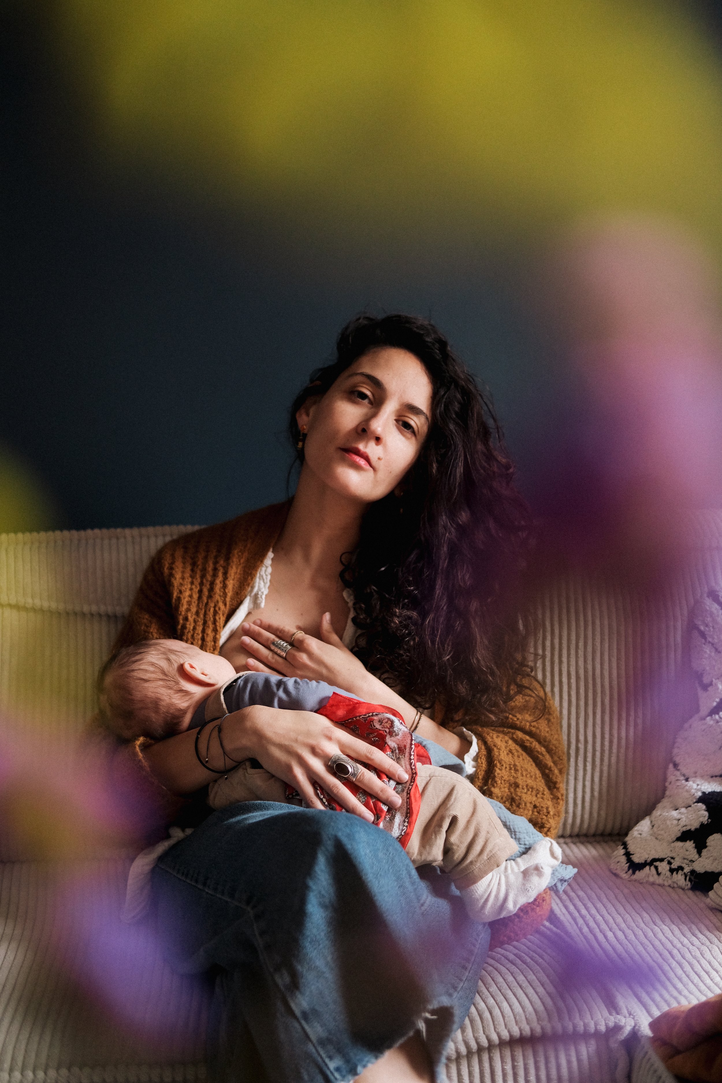 Une femme aux cheveux bouclés et longs, portant un pull marron, est assise sur un canapé, tenant un bébé dans ses bras. Le bébé est habillé en vêtements colorés et semble dormir ou être calme. La femme regarde vers l'objectif avec une expression douce.