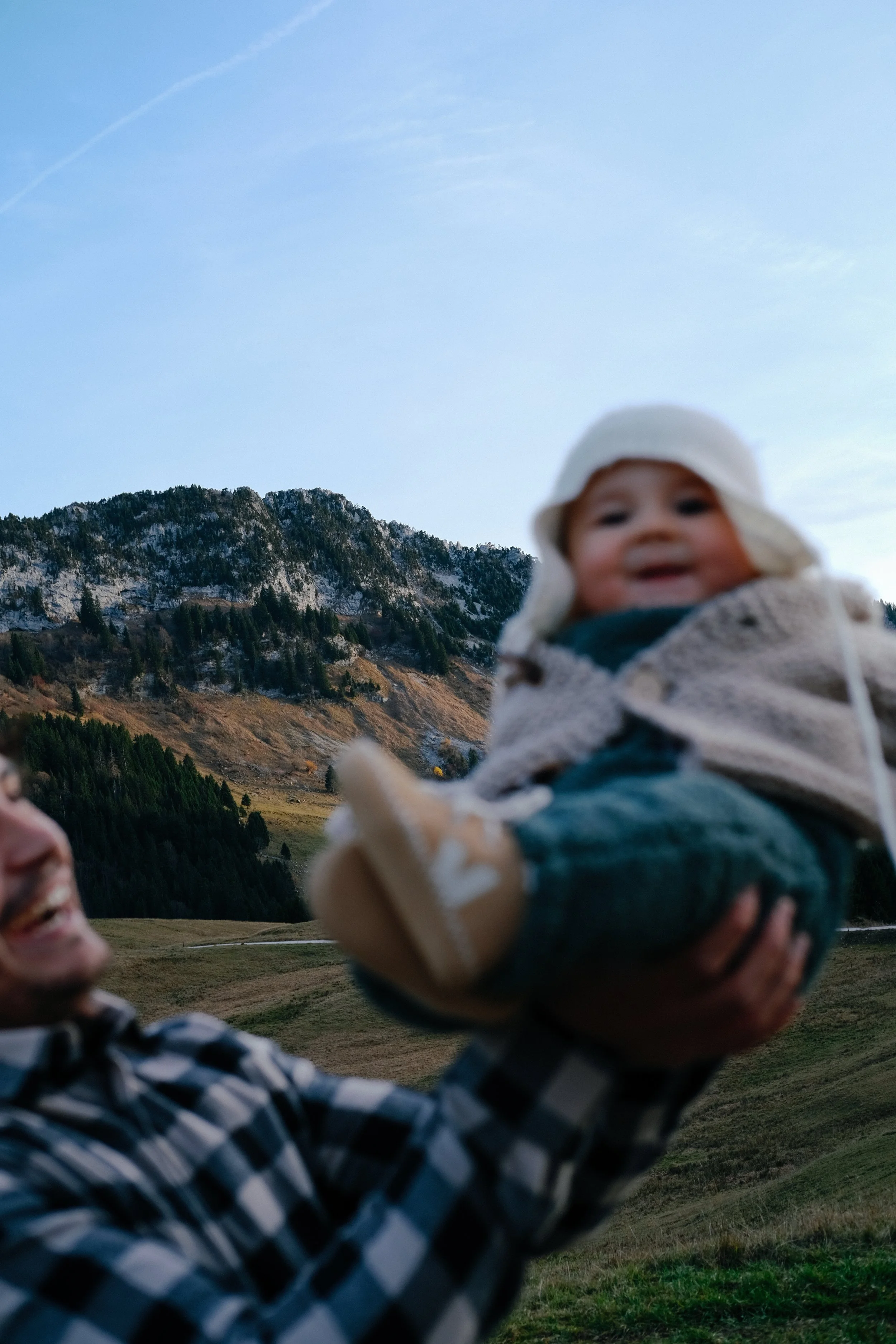 Une personne souriante tenant un bébé dans un paysage de montagnes avec des sapins et un ciel bleu clair.