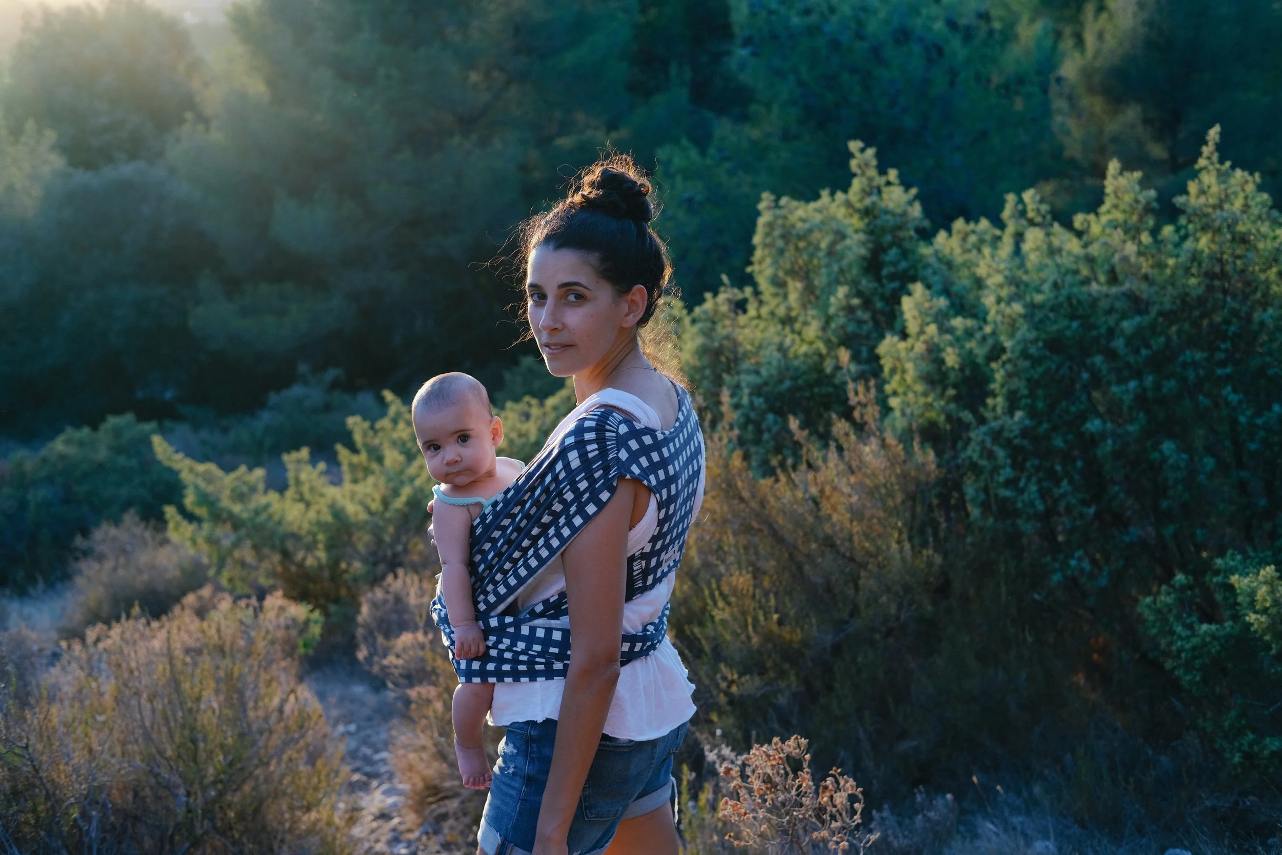 Une femme portant un bébé en portage sur le devant, dans un environnement naturel avec des arbres et des broussailles, en fin d'après-midi ou au début de soirée.