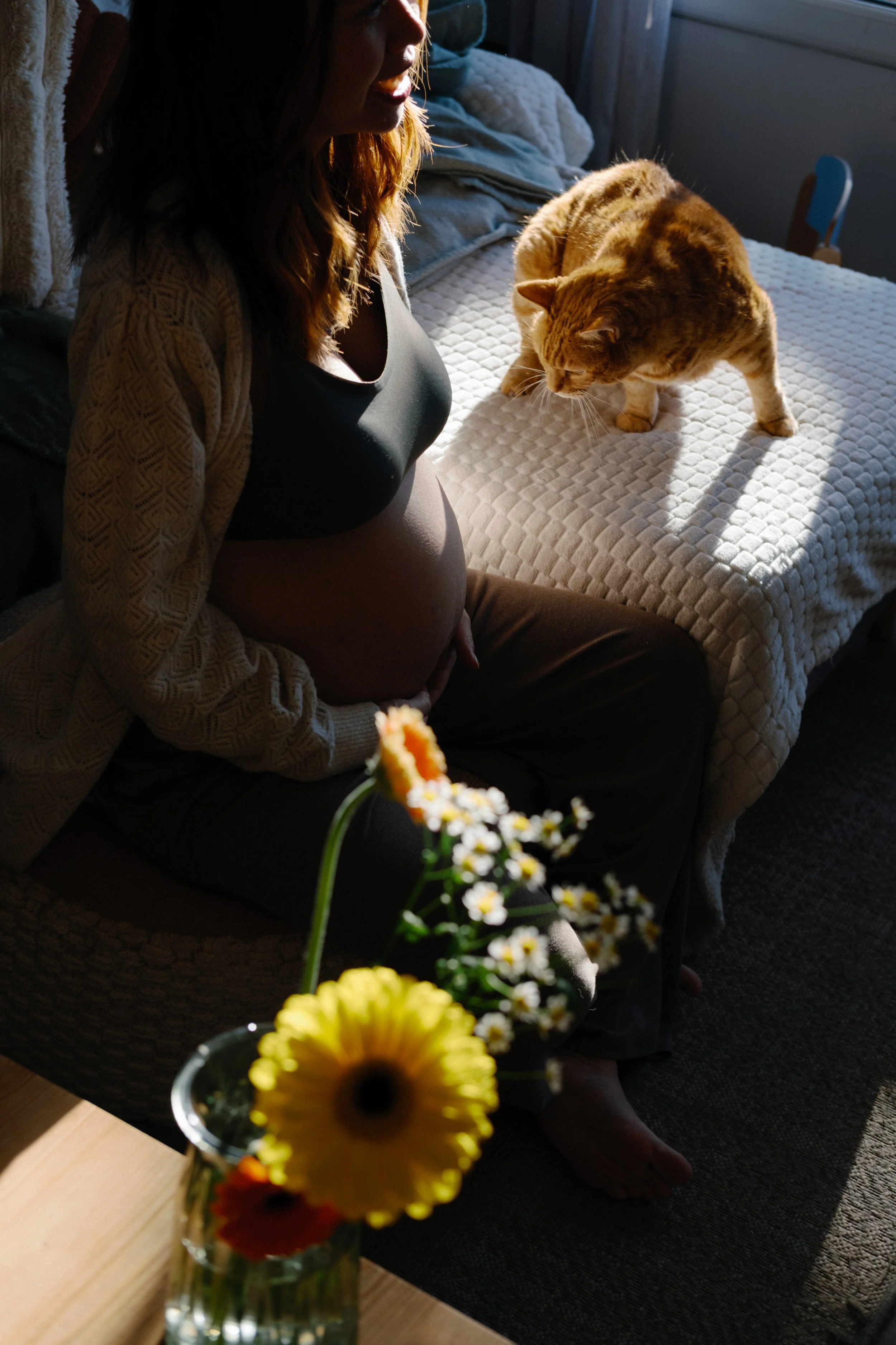 Une femme enceinte assise sur un canapé, regardant un chat orange sur un lit à côté d'elle.