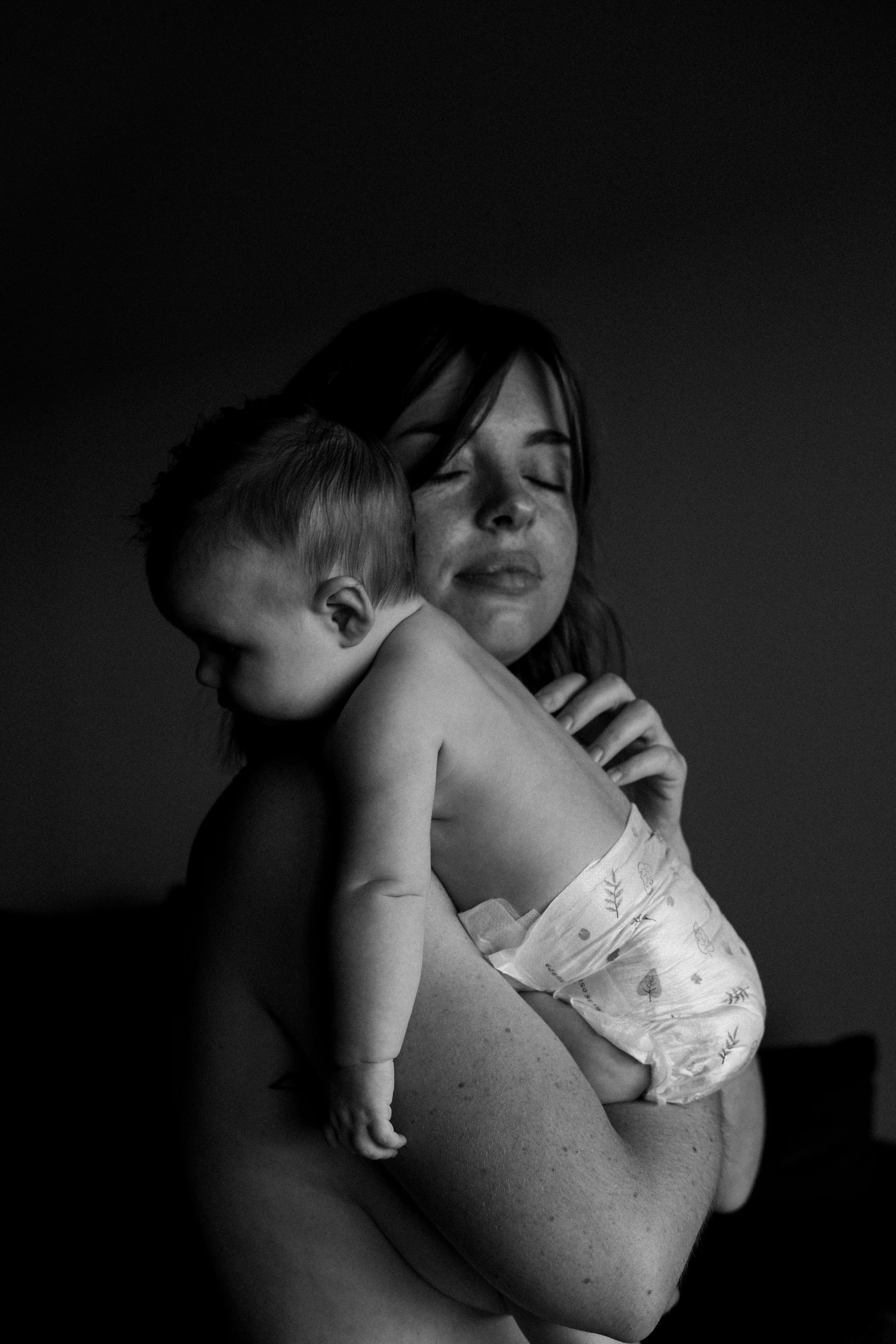 Une femme allaitante tenant un bébé contre son épaule, en toile de fond sombre.