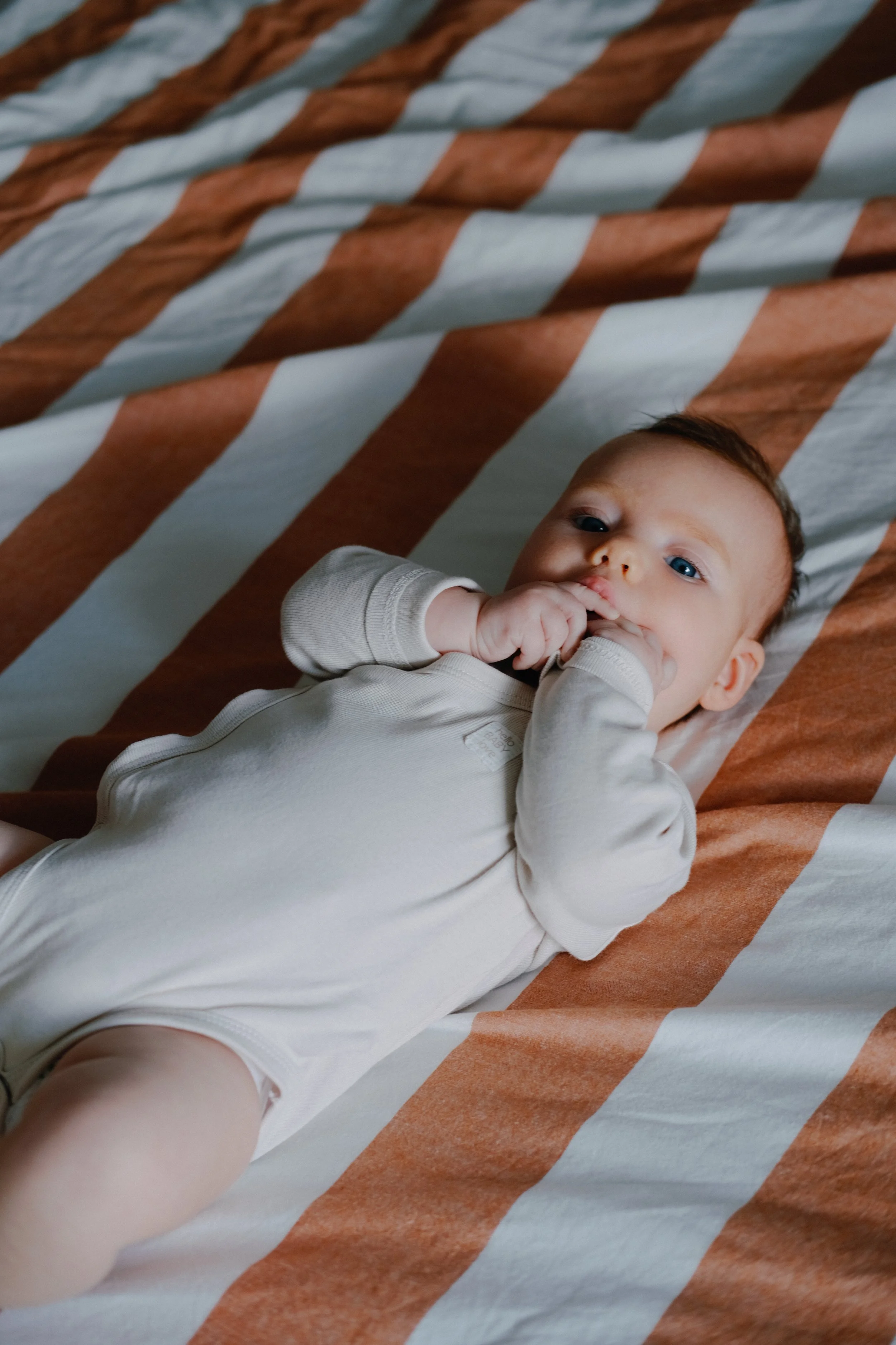 Un bébé allongé sur un lit avec des draps rayés beige et rouille. Le bébé porte une tenue beige clair et a les yeux bleus, avec les mains près du visage.
